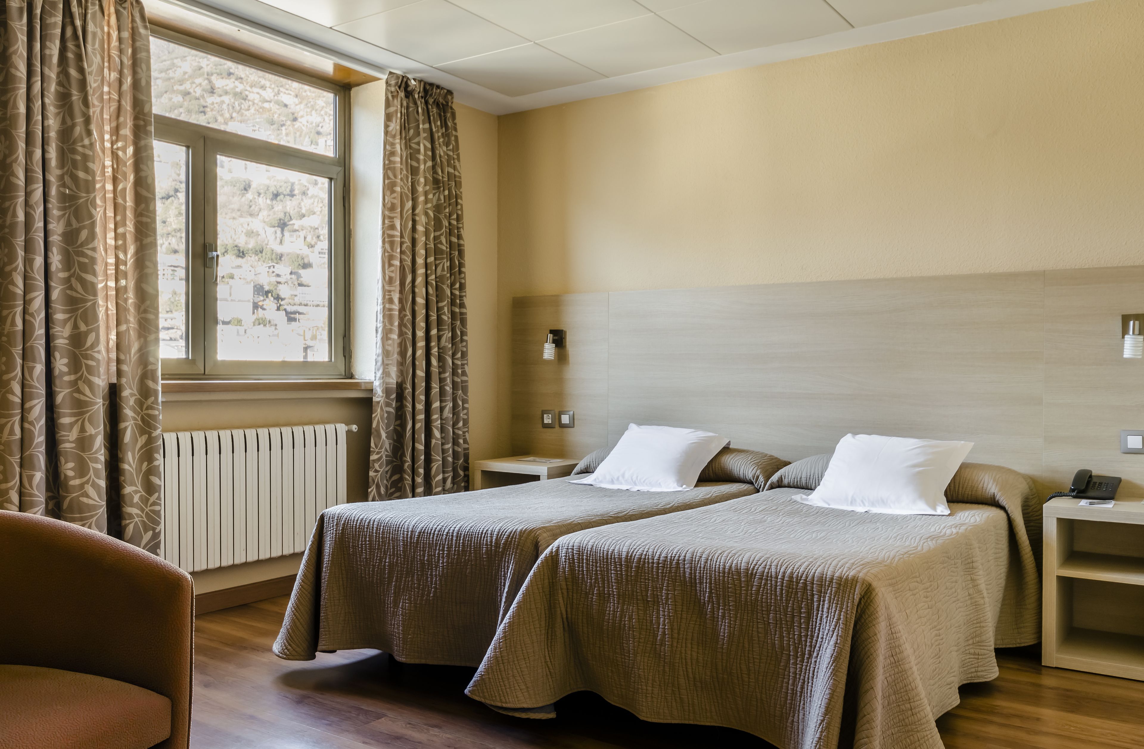 A hotel room with two beds, a window with curtains, a bedside table with a lamp, and a chair.