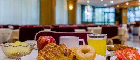 Un desayuno con cruasanes, un donut, un vaso de jugo de naranja, un plato de rebanadas de manzana y una taza, además de un plato con rodajas de tomate.