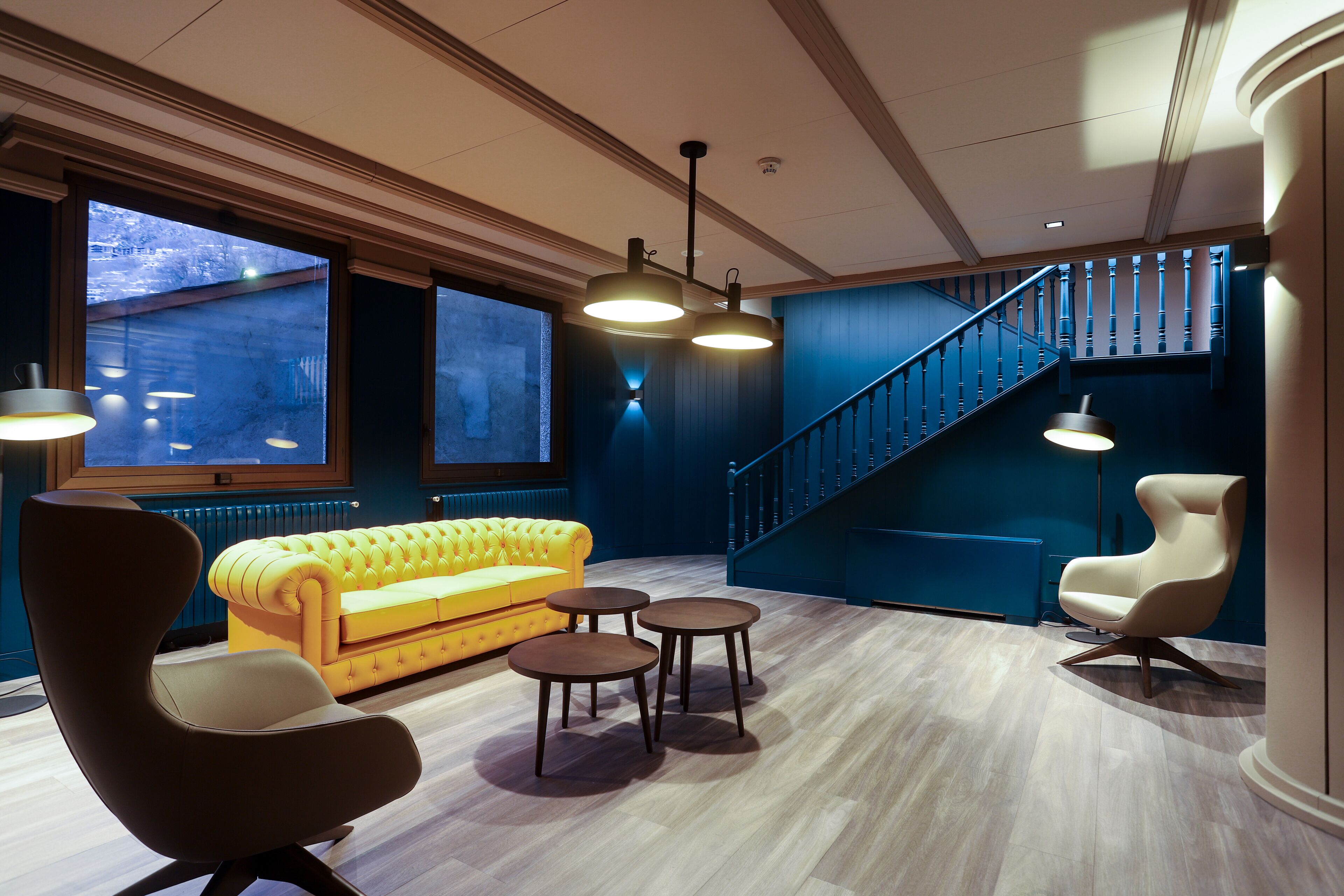 A modern interior with a yellow sofa, a brown armchair, and a white chair. There are two small tables and a staircase with blue walls.