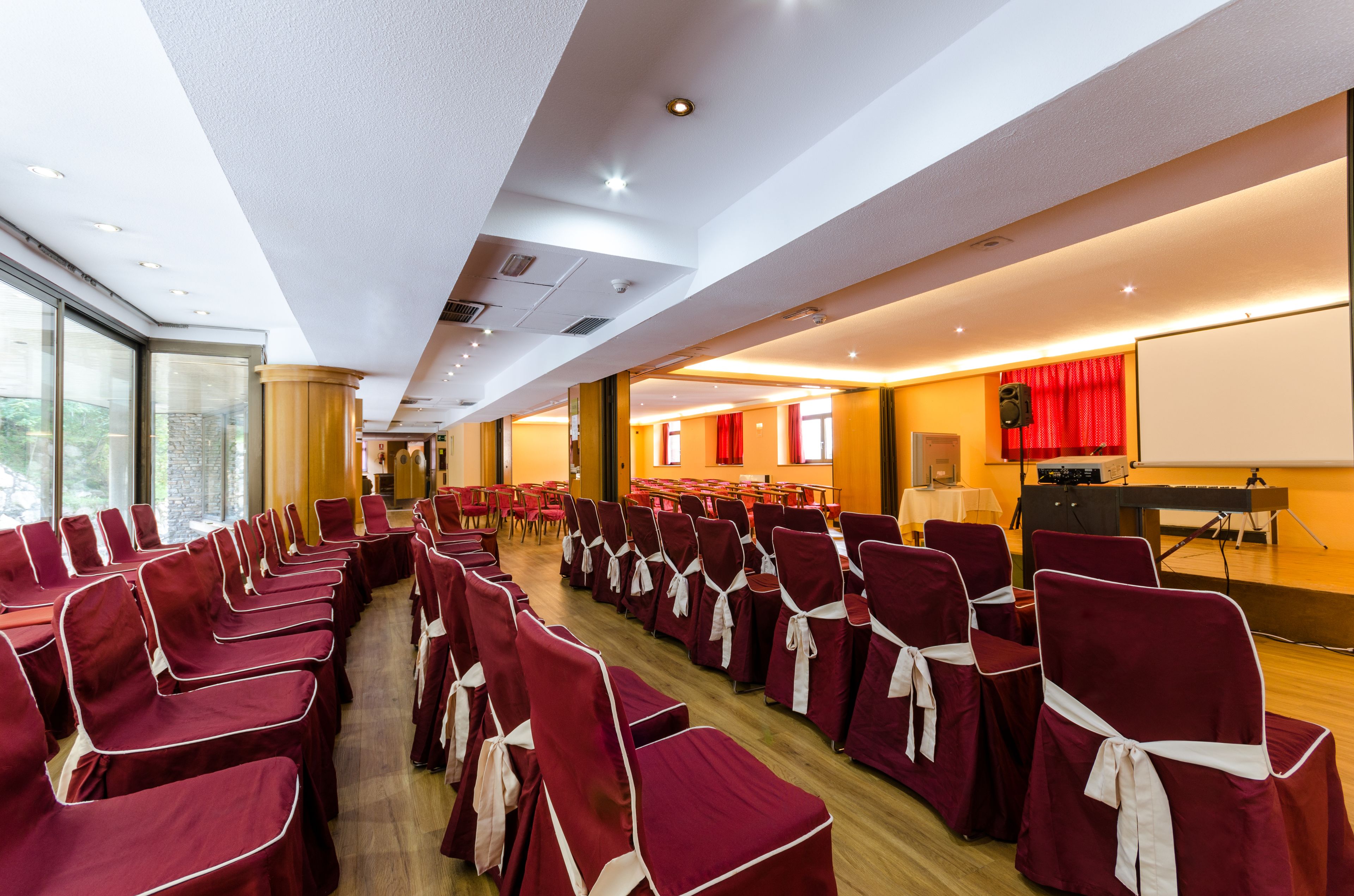 A conference room with rows of chairs, a stage with a projector, and a large window providing natural light.