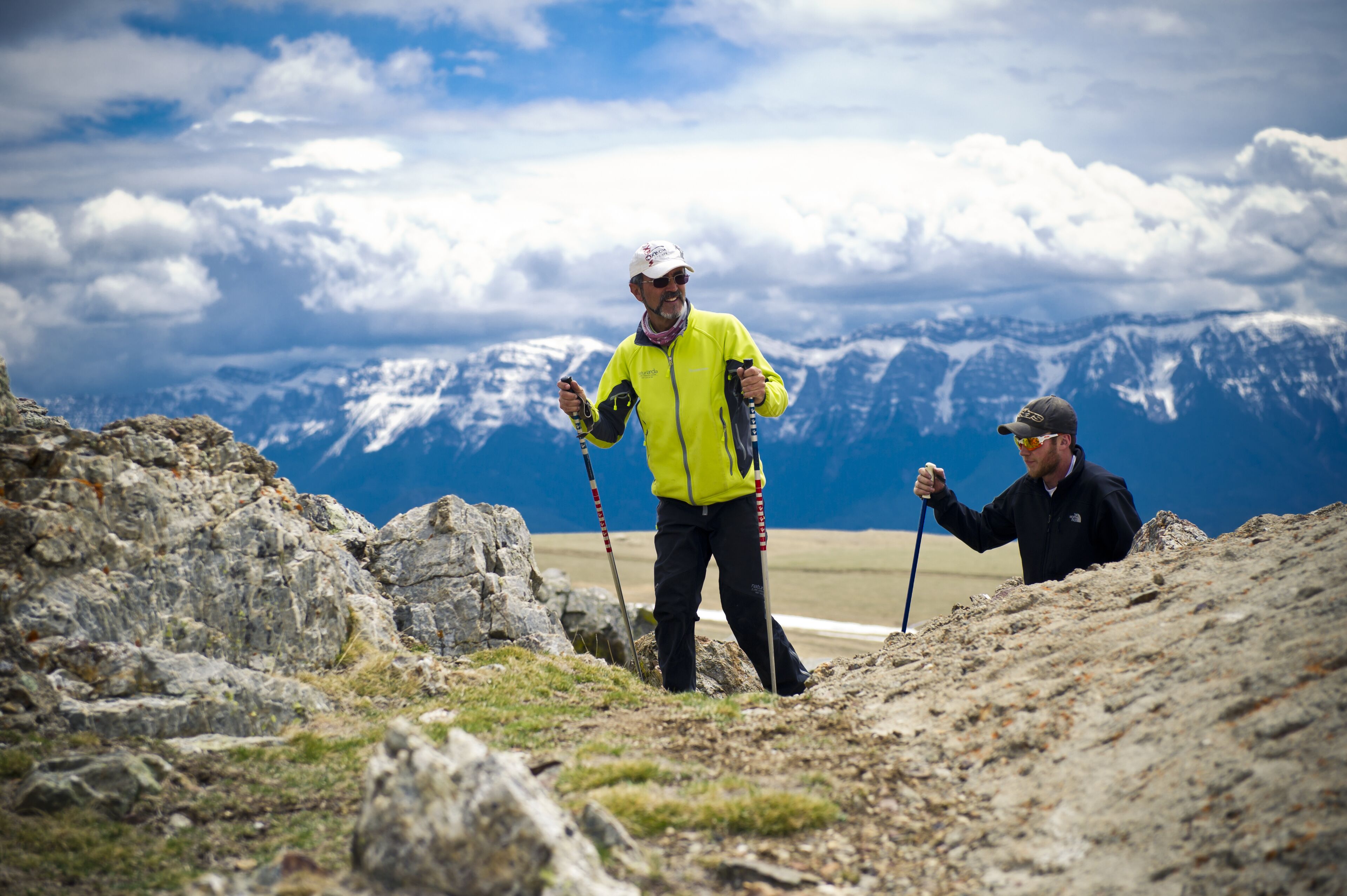 Deux randonneurs sur un sentier de montagne rocailleux, avec en arrière-plan une vue sur des montagnes enneigées.