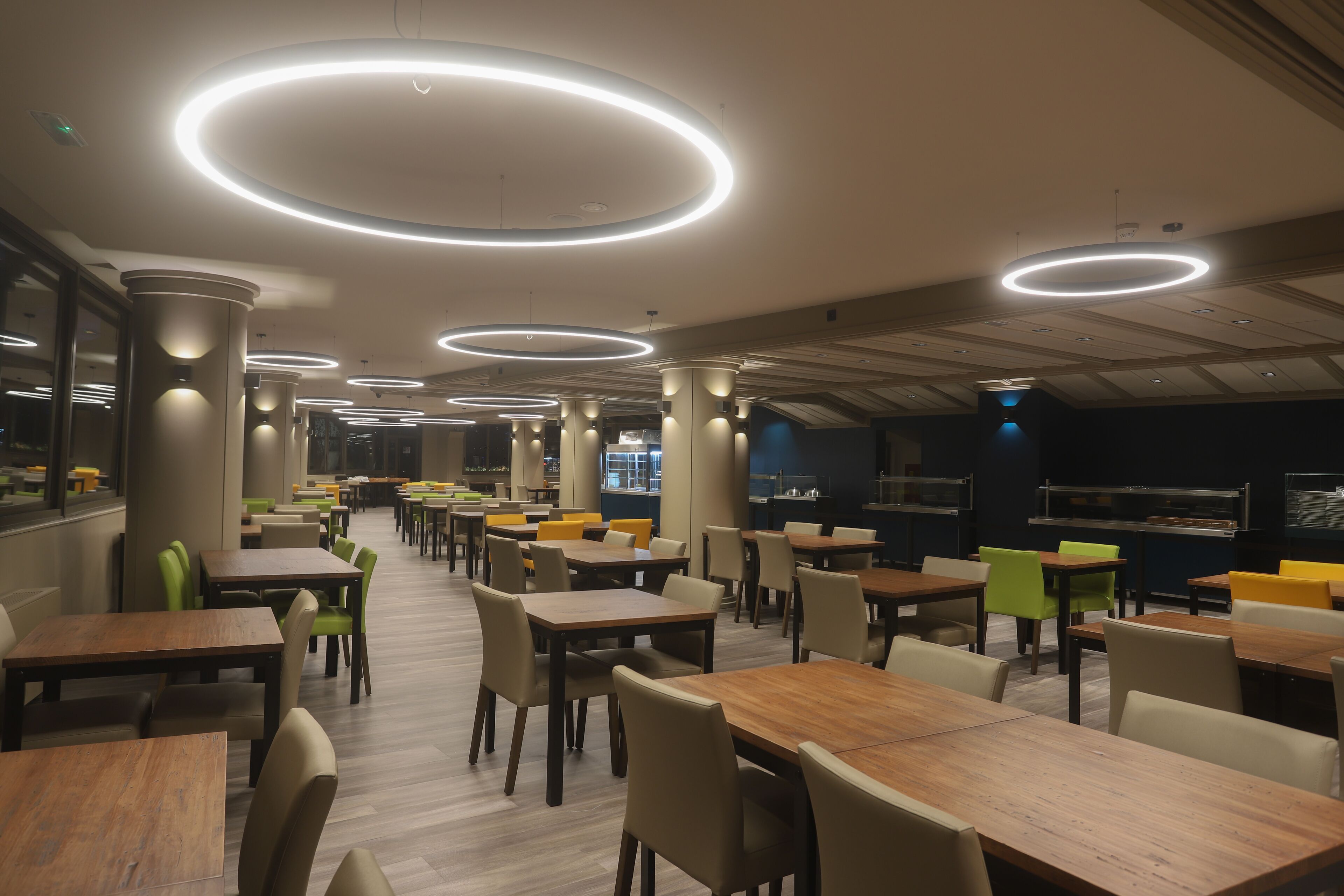Um restaurante espaçoso com várias mesas e cadeiras, luminárias de teto circulares e um design de interiores moderno.