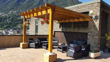 Une terrasse sur le toit, aménagée avec du mobilier d’extérieur, une pergola ornée de suspensions fleuries et un bâtiment en pierre offrant une vue sur un paysage montagneux.