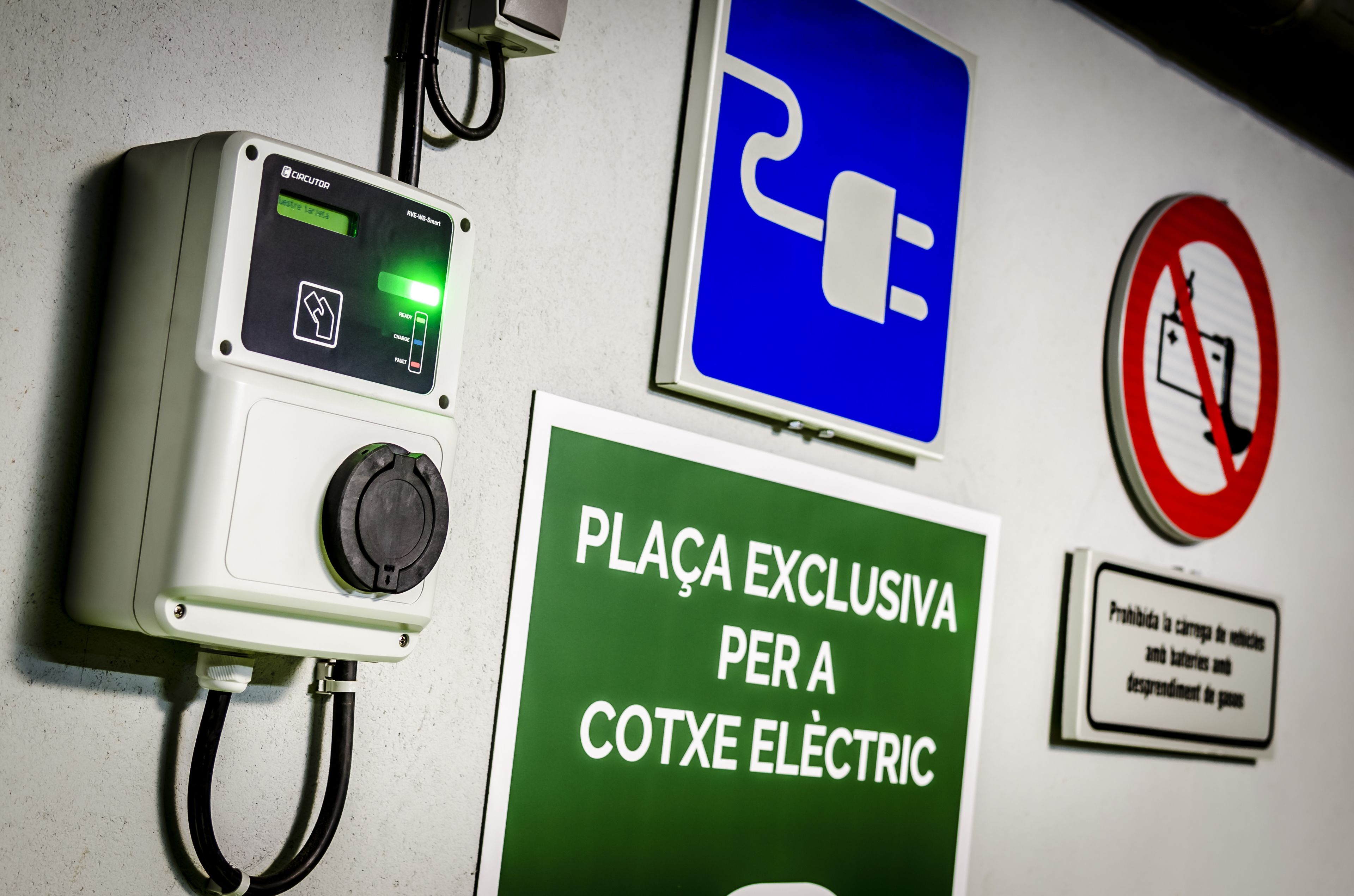 Une boîte électrique murale dotée d’un écran numérique, d’un symbole de prise de couleur bleue et d’un panneau vert indiquant une zone de stationnement réservée aux véhicules électriques.