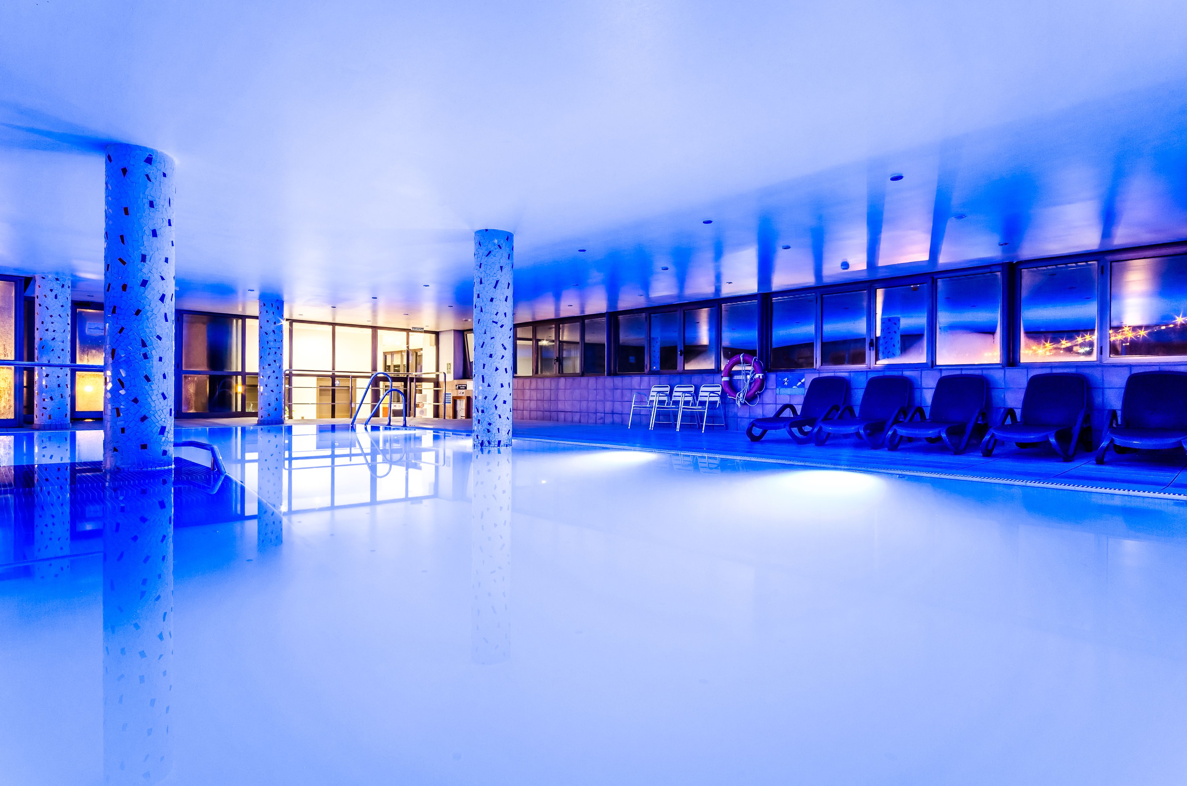 Una piscina interior moderna con paredes de vidrio transparente, un área de estar con sillones y una columna central.