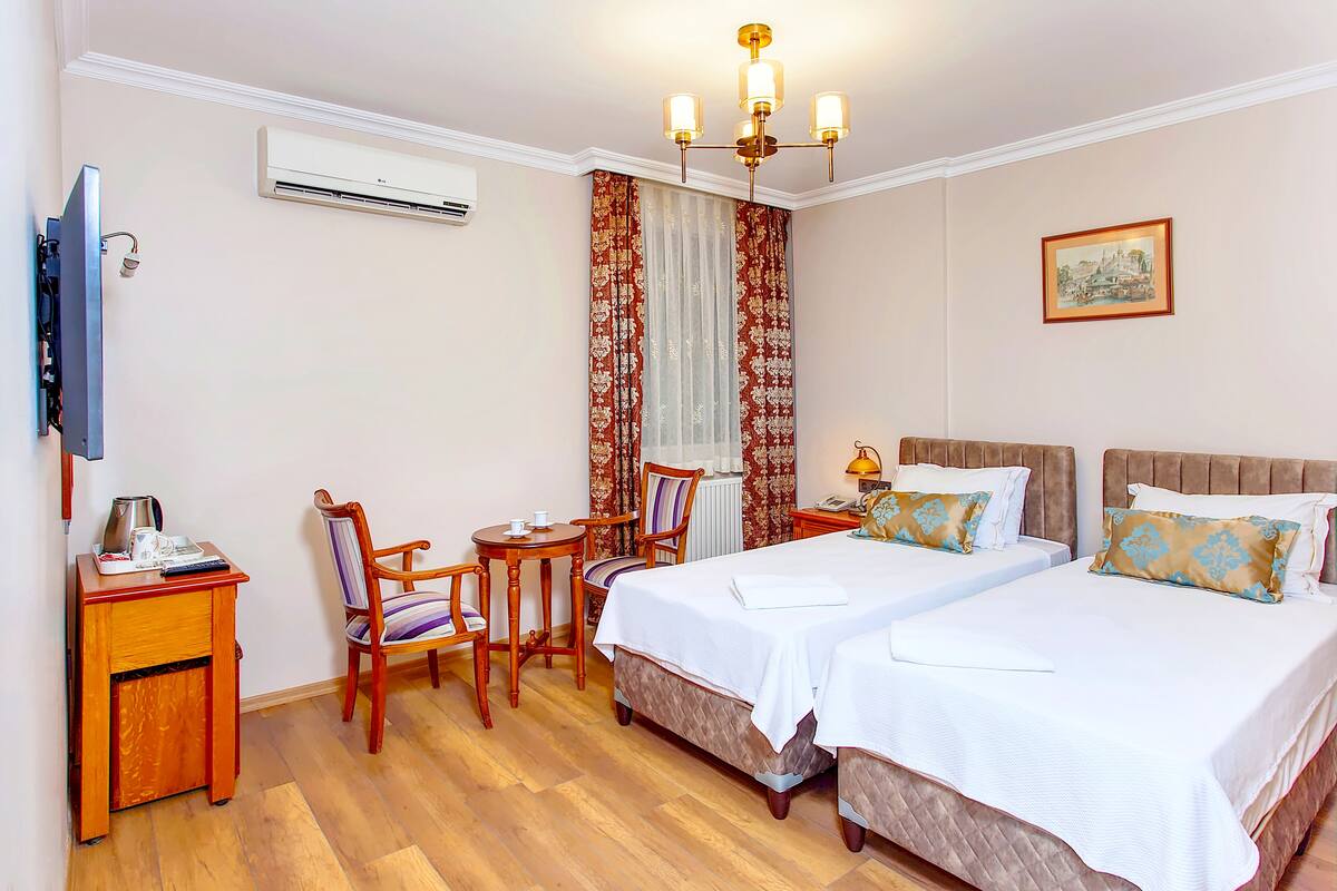 A hotel room with two beds, a desk, a chair, a TV, an air conditioner, and a chandelier.