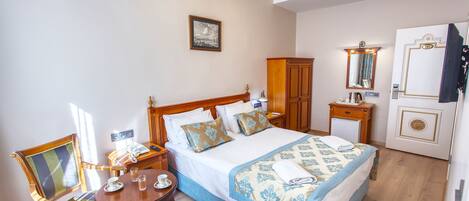 Quarto de hotel com cama, uma cadeira de madeira, uma pequena mesa com uma chávena e copos, mesa de cabeceira, guarda-roupa, espelho e uma porta com um casaco pendurado.