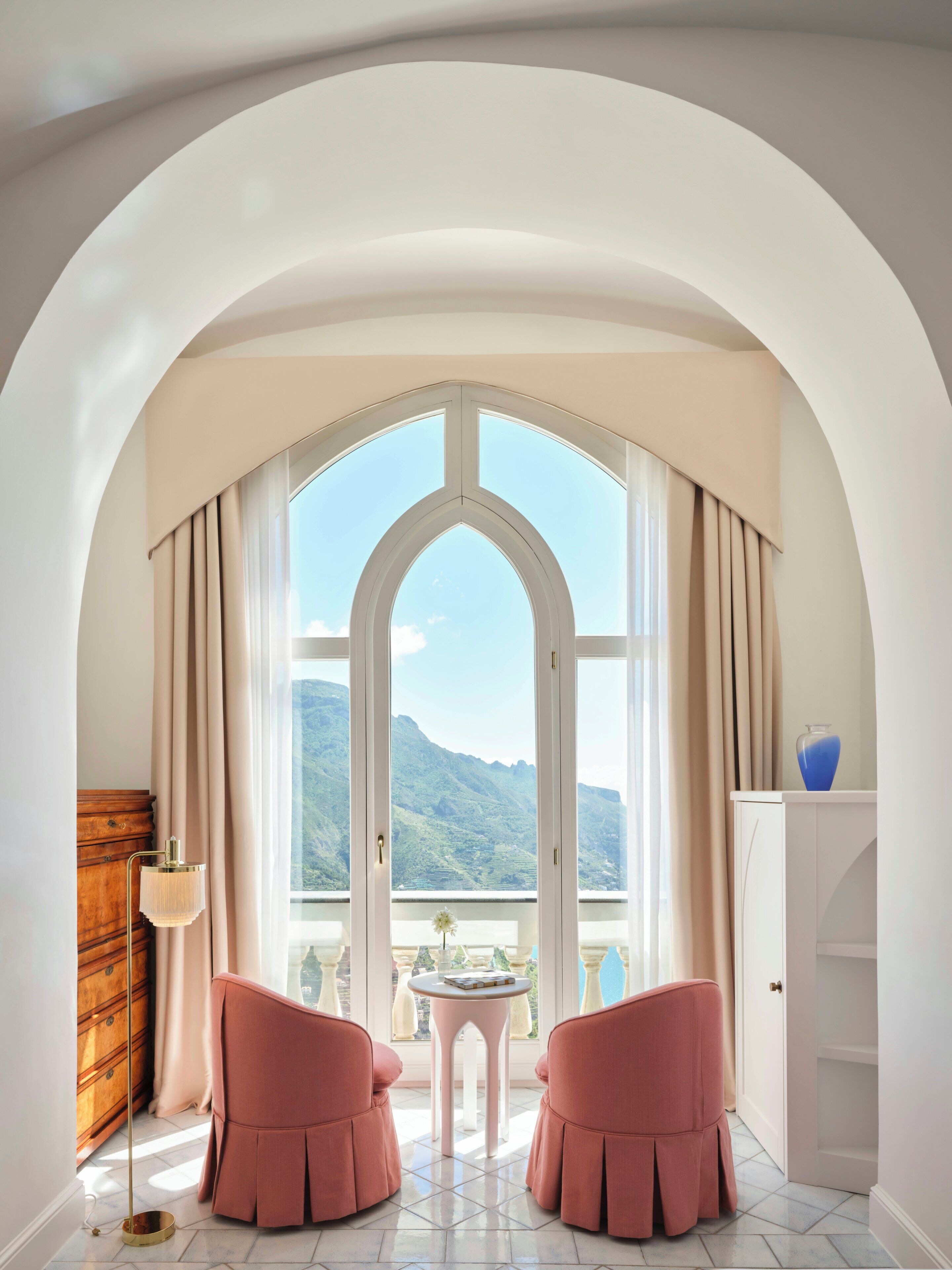 Una stanza con un'ampia finestra ad arco, due poltrone rosa, un tavolino rotondo e un comò in legno.