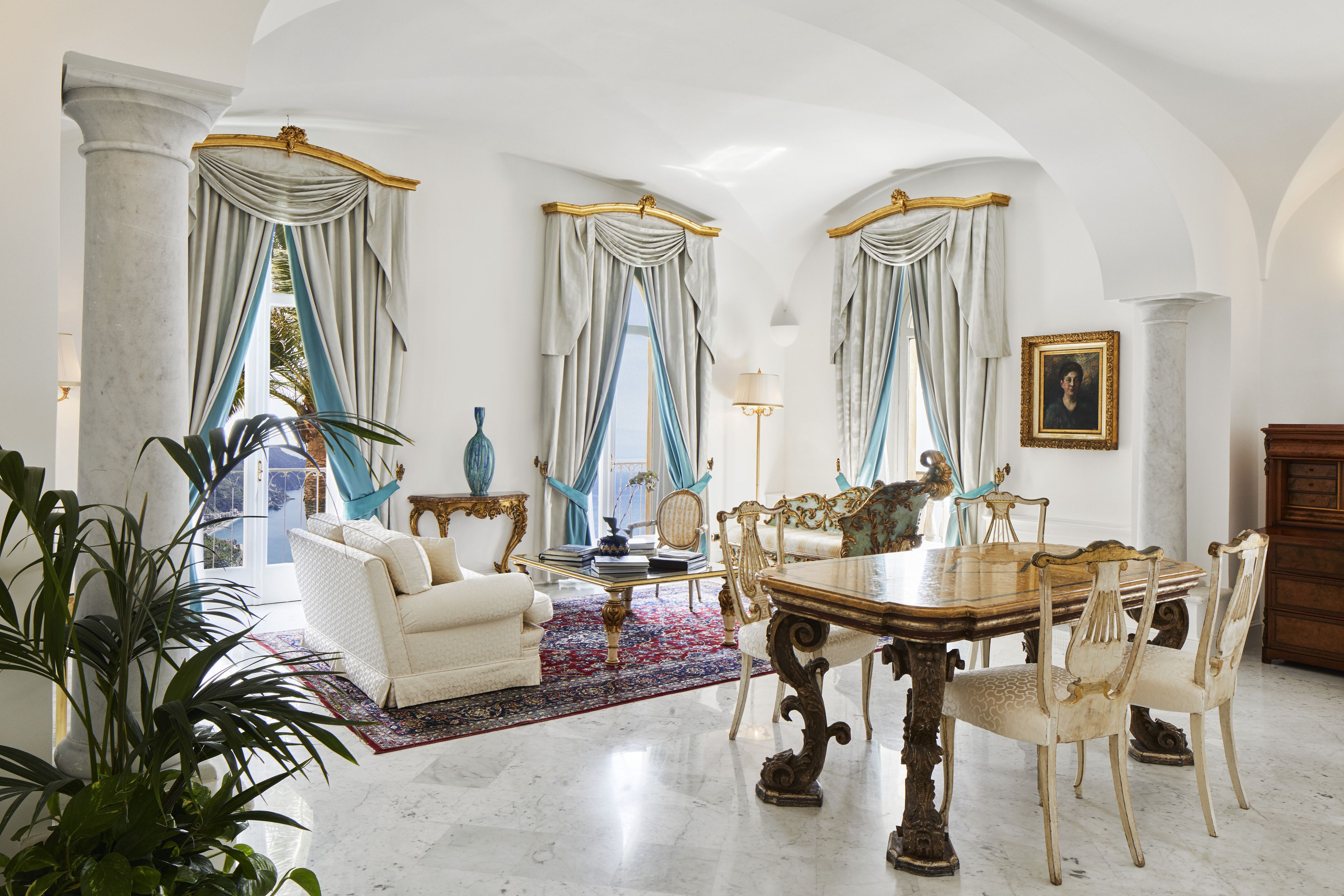 A spacious room with a marble floor, a white sofa, a dining table with chairs, and a large window with curtains.