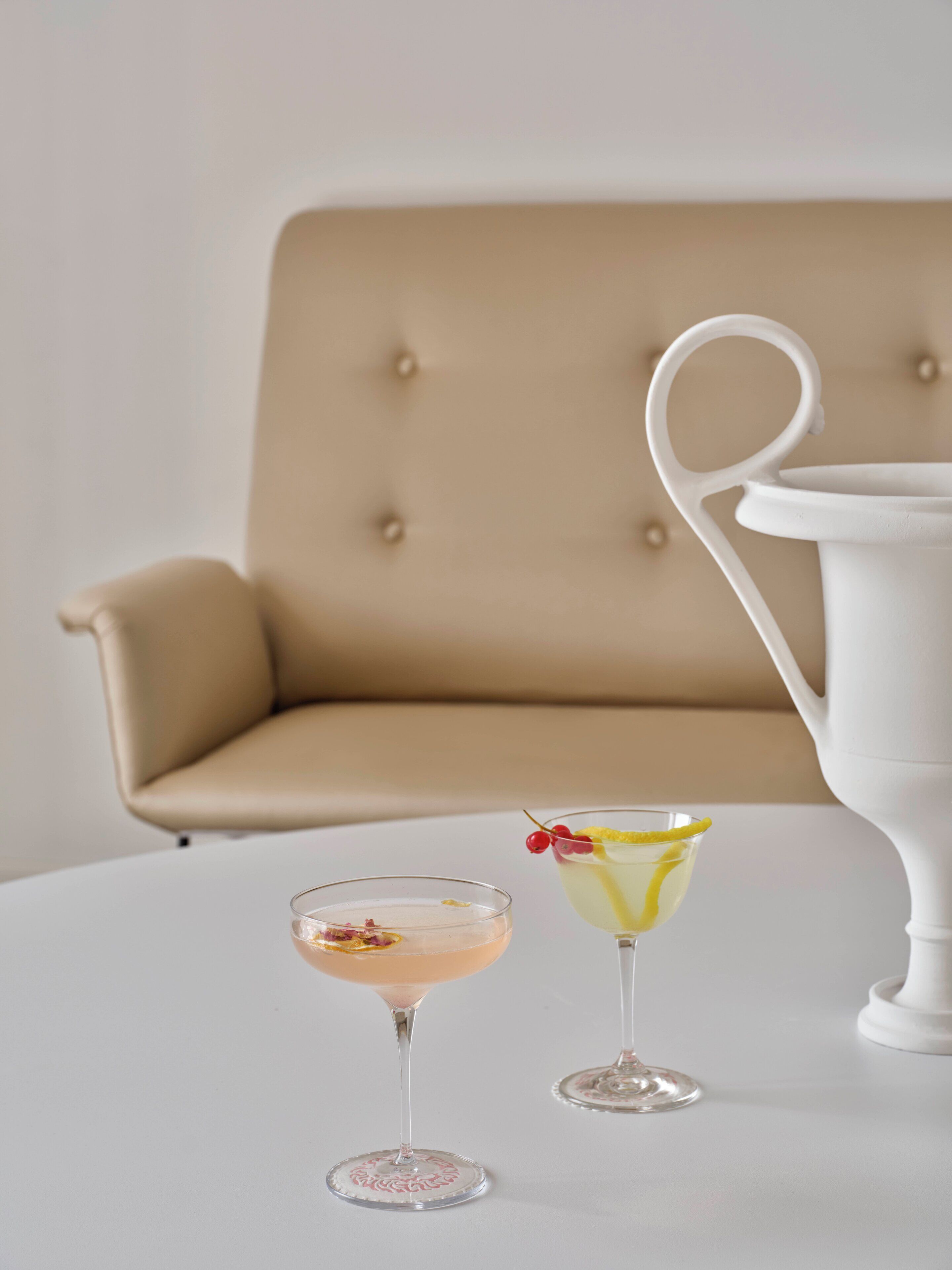 Two cocktails on a white table with a beige upholstered chair and a white pitcher in the background.