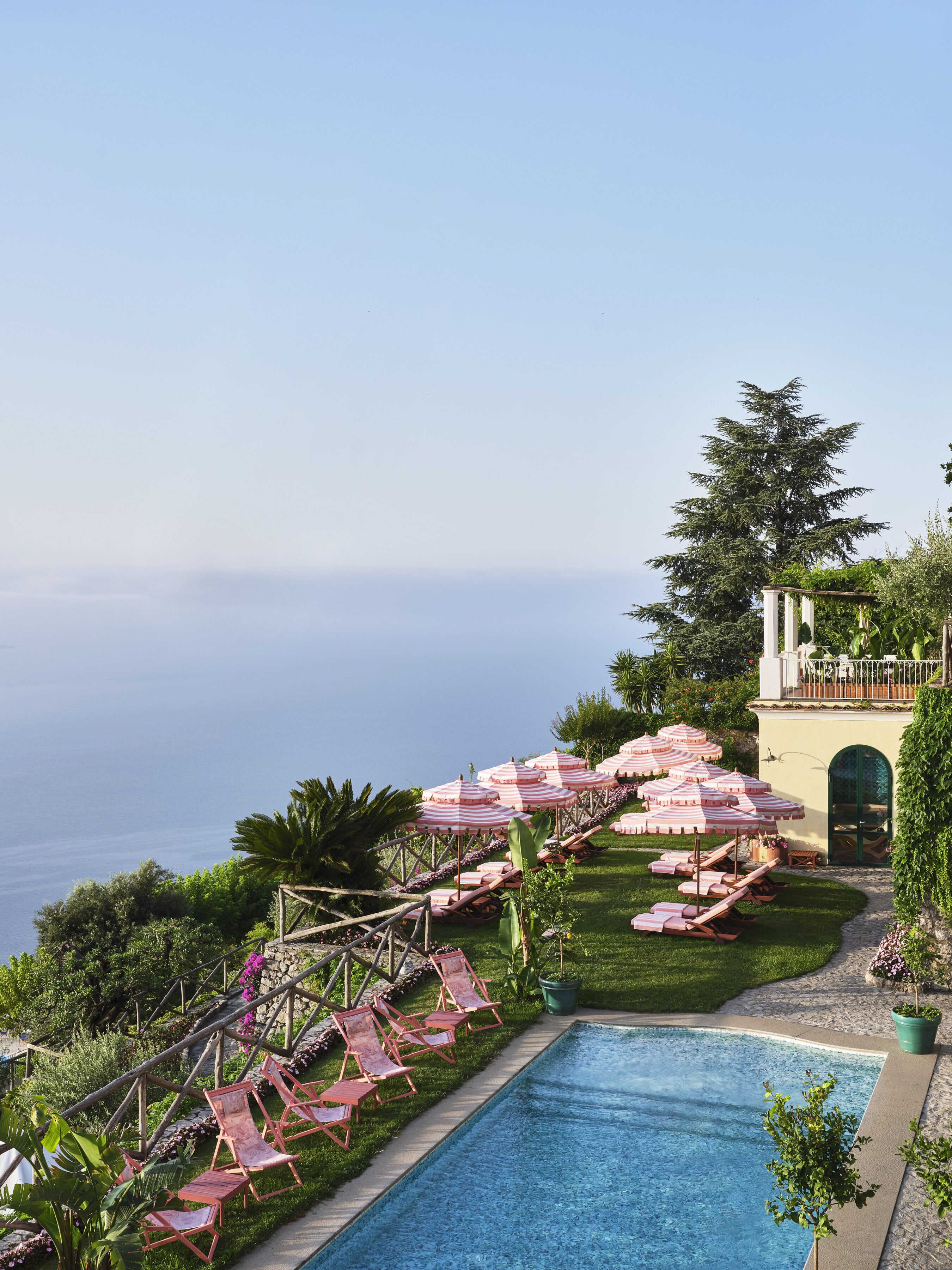 Un espace aménagé au bord de la piscine, avec des chaises longues et des parasols, une vue dégagée sur l’océan et un bâtiment doté d’une entrée en arc de cercle.