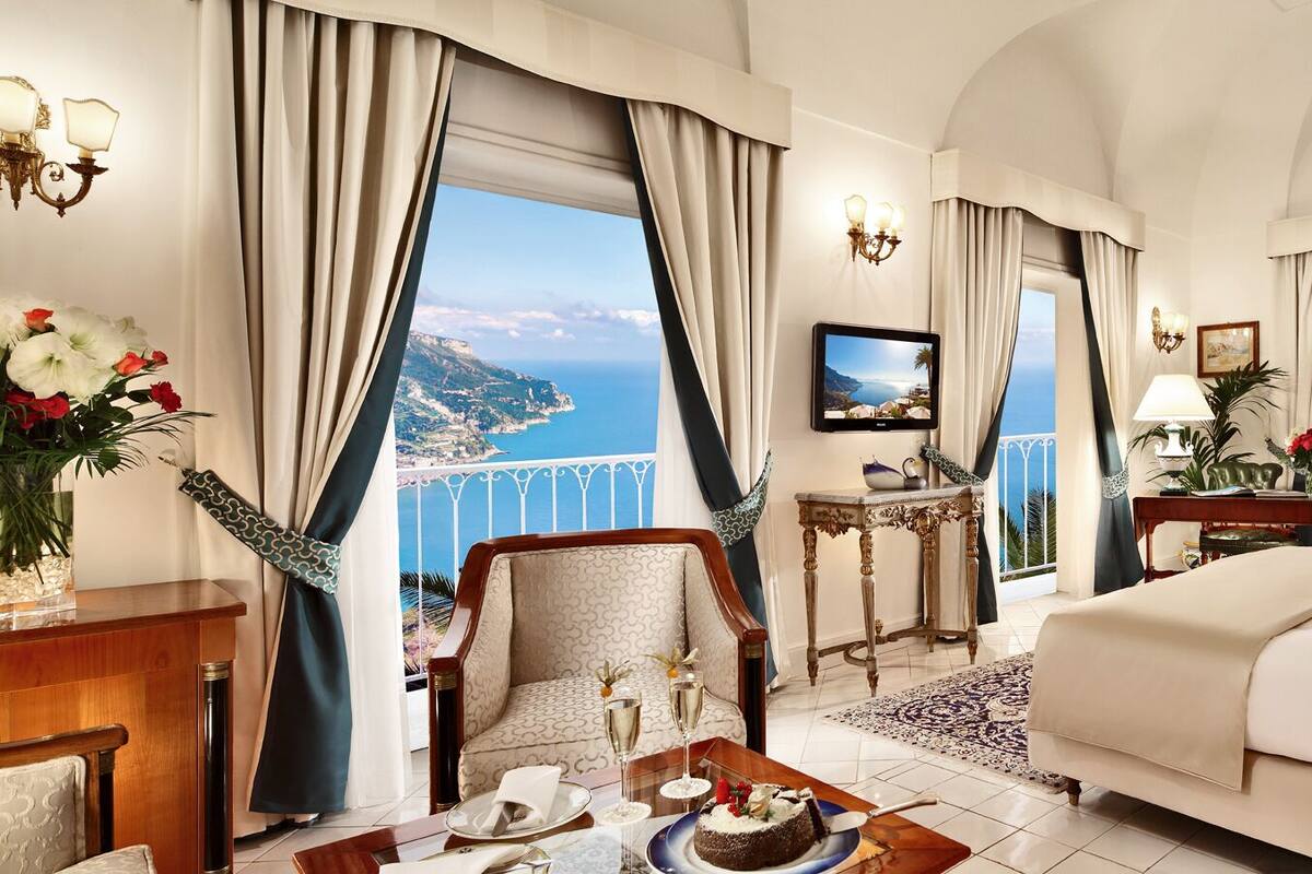 Una lujosa habitación de hotel con una cama grande, una zona de estar con vista al mar y un balcón con cortinas.