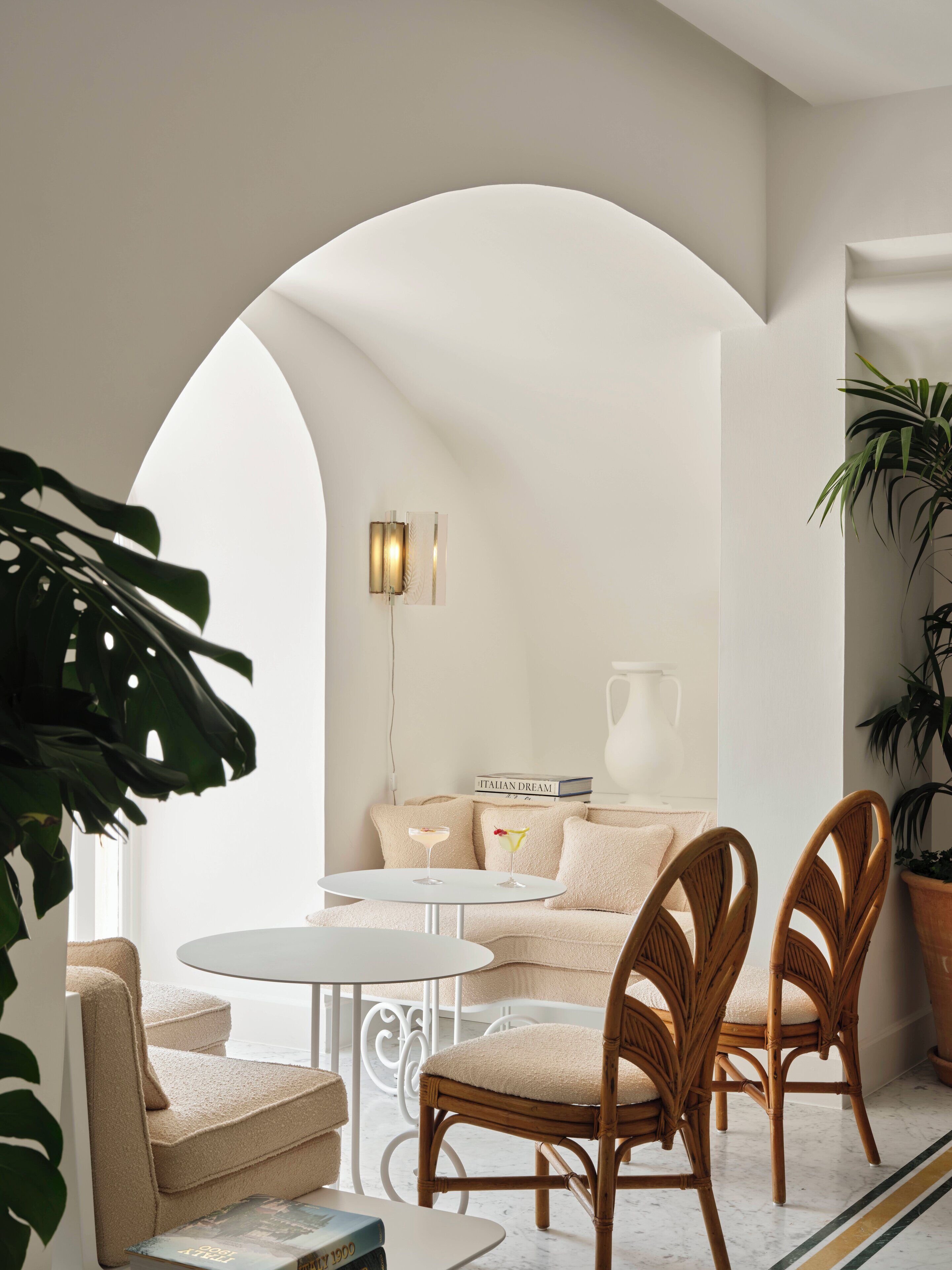Un interior acogedor con una mesa redonda, dos sillas de mimbre, un sofá con cojines, una planta en maceta y una lámpara de pared.