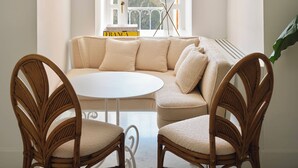 A room with a white arched window, a round table, two wicker chairs, a beige sofa, and a view of a mountainous landscape.