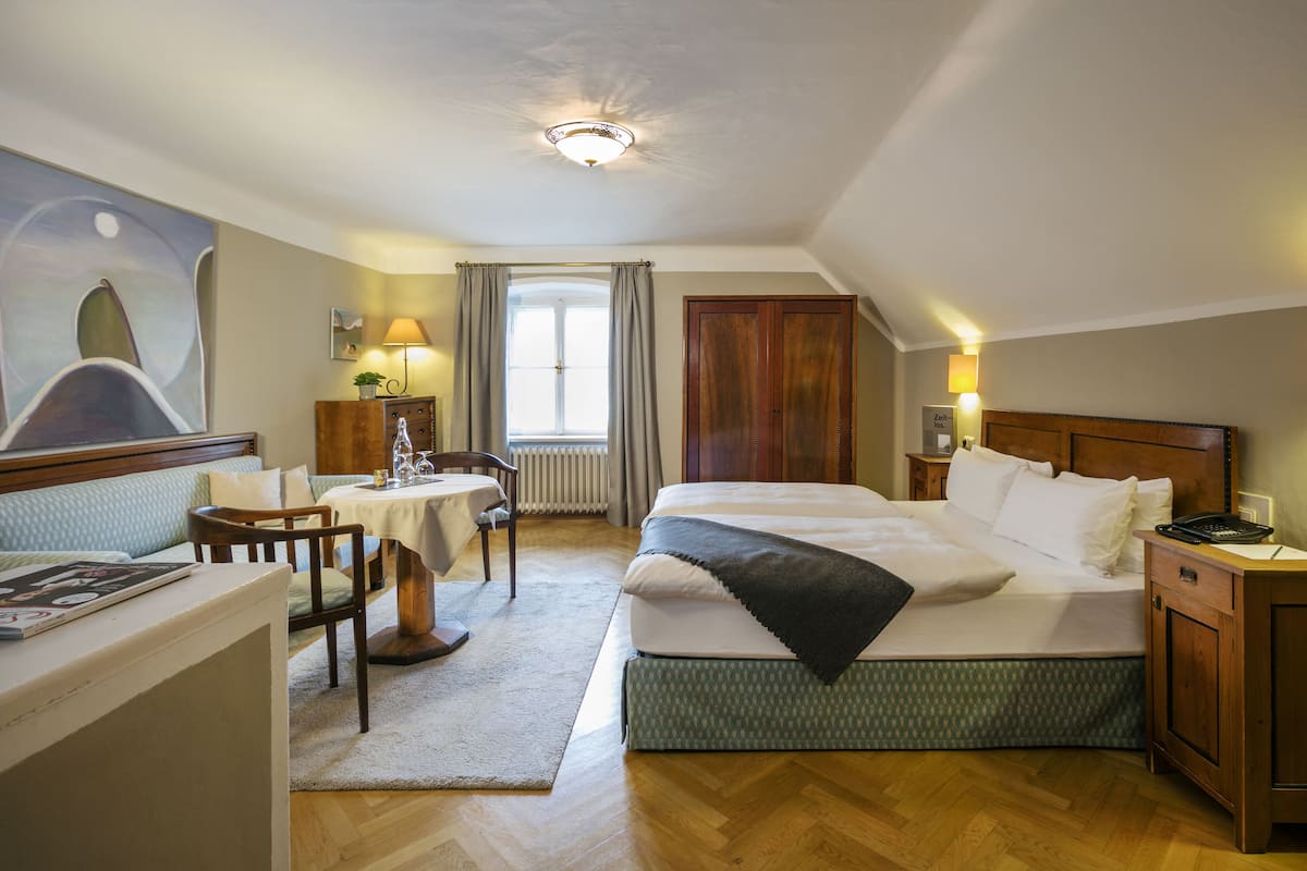 Une chambre d’hôtel avec un lit, un bureau, une chaise, une table, une lampe, une fenêtre avec des rideaux et un tableau au mur.