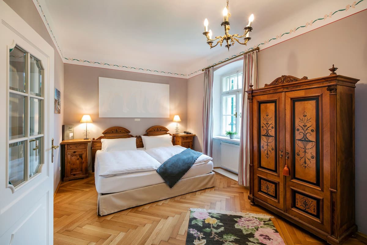 Une chambre à coucher avec un lit double, deux tables de chevet avec des lampes, une armoire en bois, un lustre et une fenêtre avec des rideaux.