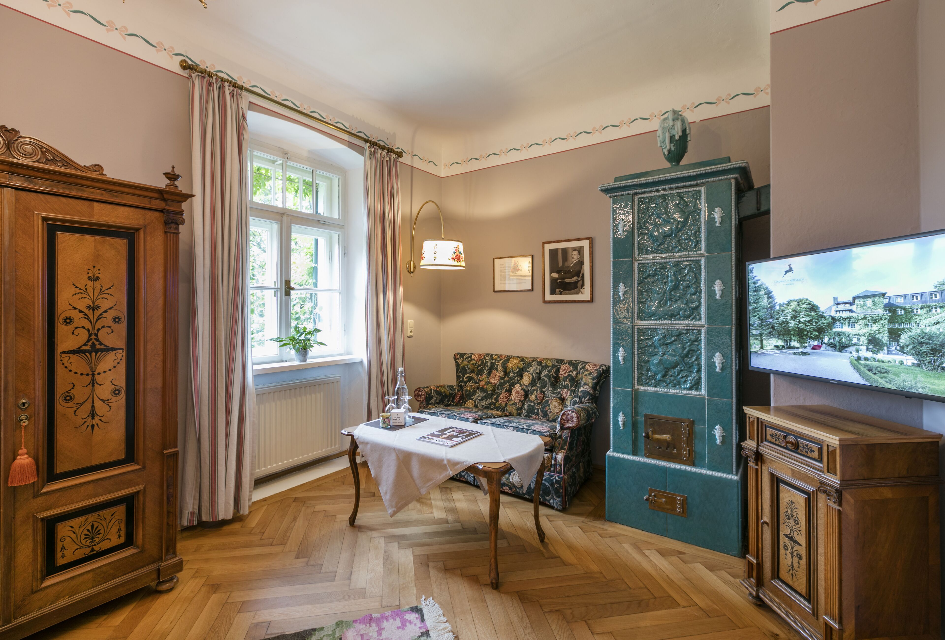 Una habitación con piso de madera, una mesa con mantel blanco, un sofá floral, un aparador con puertas de cristal y un televisor en la pared.