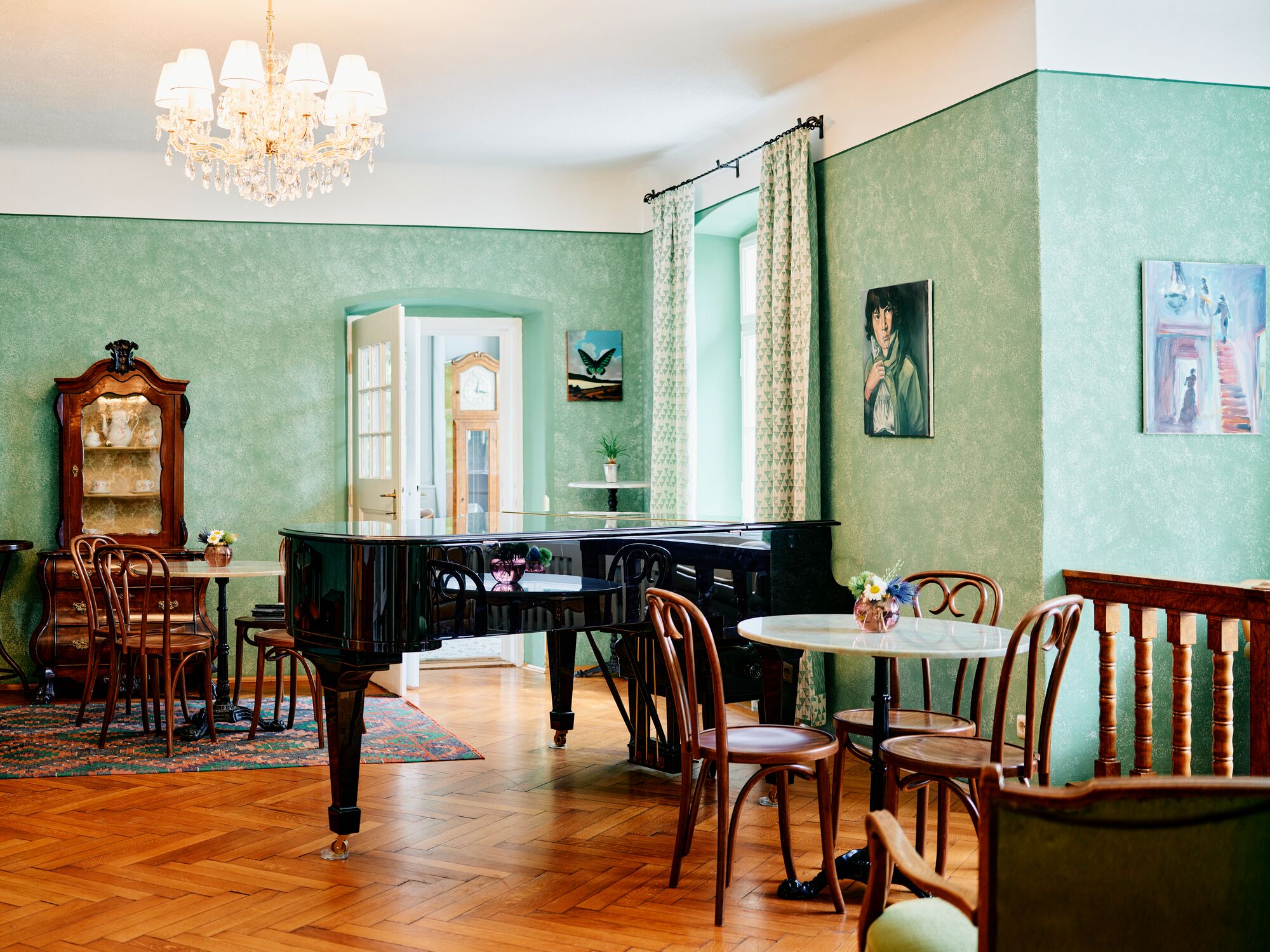 A room with a piano, wooden chairs, a chandelier, and a green wall.