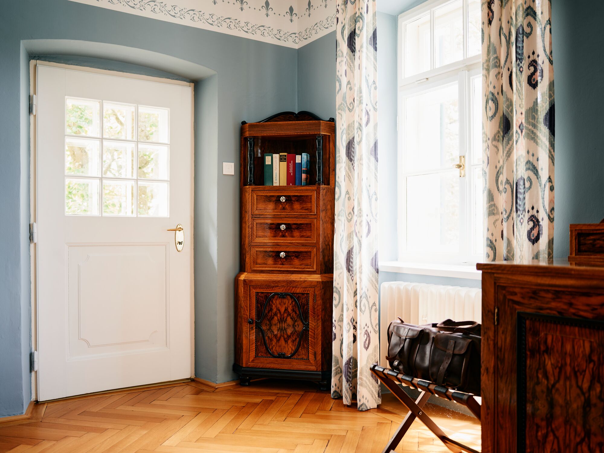 Ein Zimmer mit einem Holzkasten, einer weißen Tür und einem Fenster mit gemusterten Vorhängen.