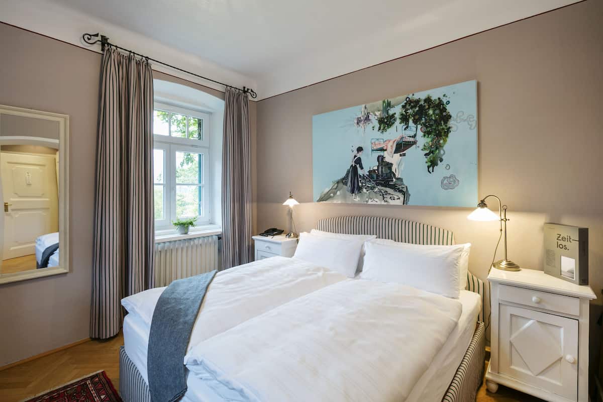 Une chambre à coucher avec un lit double, une table de chevet avec une lampe, une commode, une fenêtre avec des rideaux, un miroir et un tableau mural.