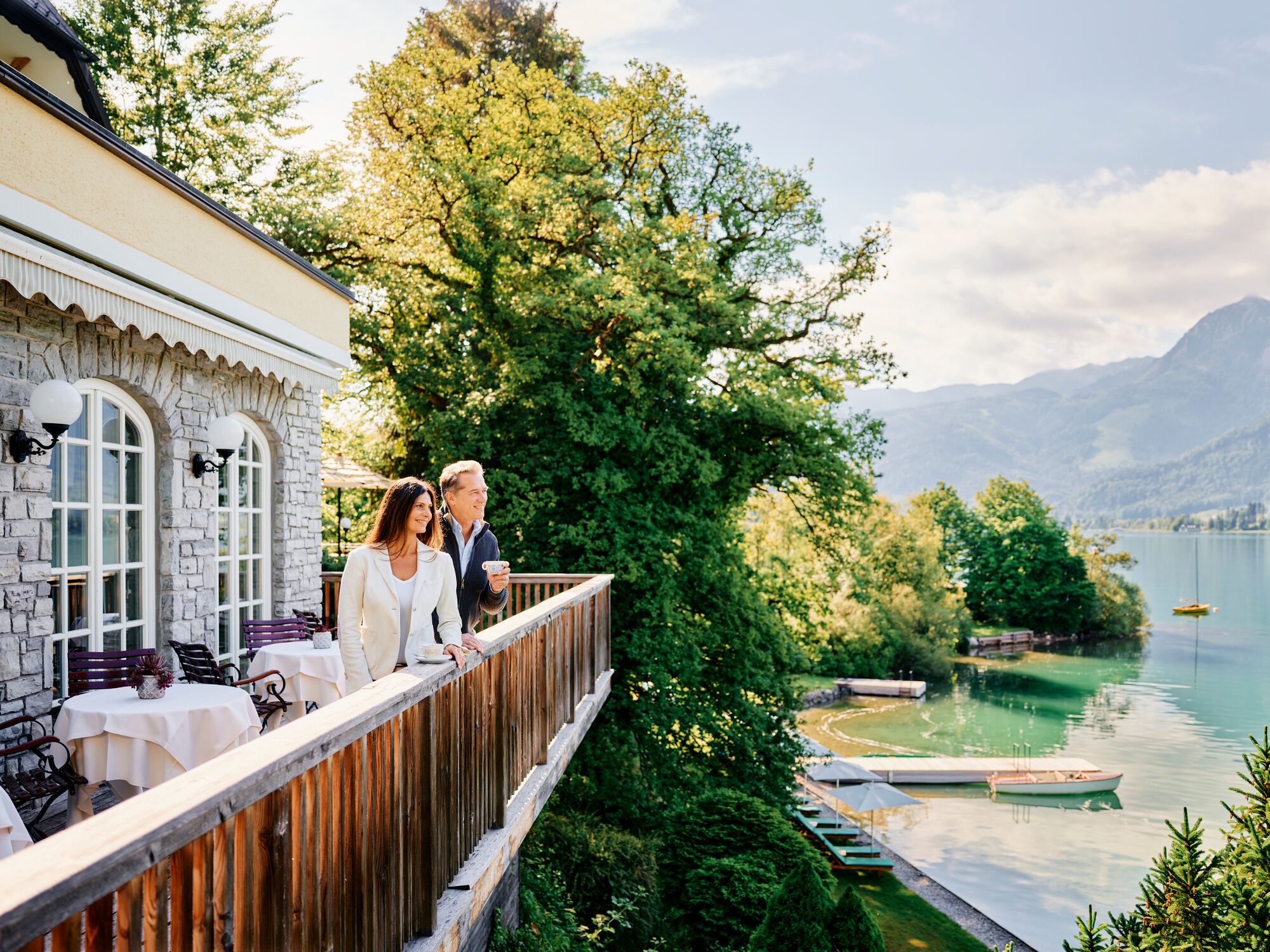 A lakeside terrace with stone walls, outdoor seating, and a view of the lake and mountains.