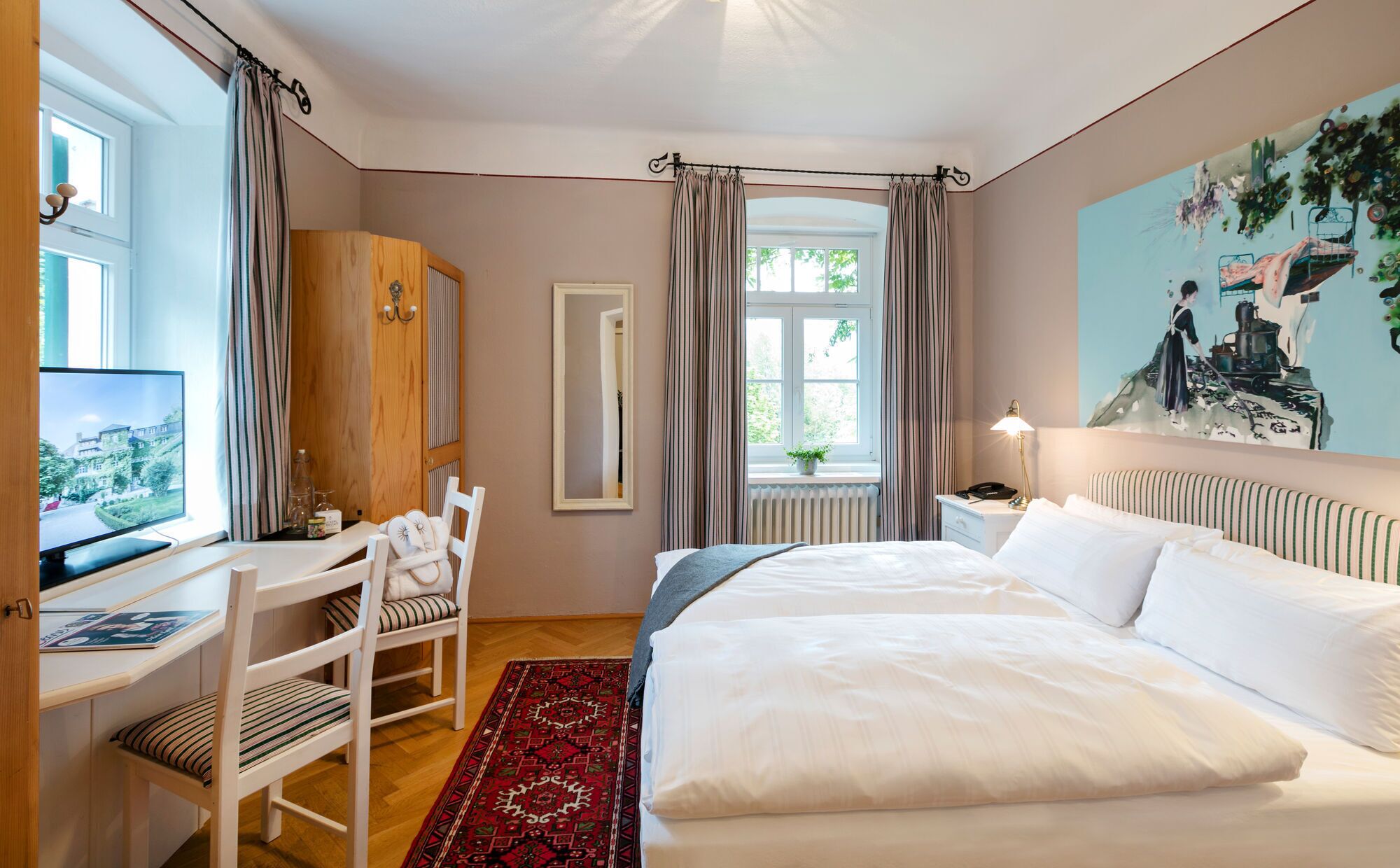 Habitación de hotel con cama, escritorio con computadora, silla, ventana con cortinas y un cuadro en la pared.