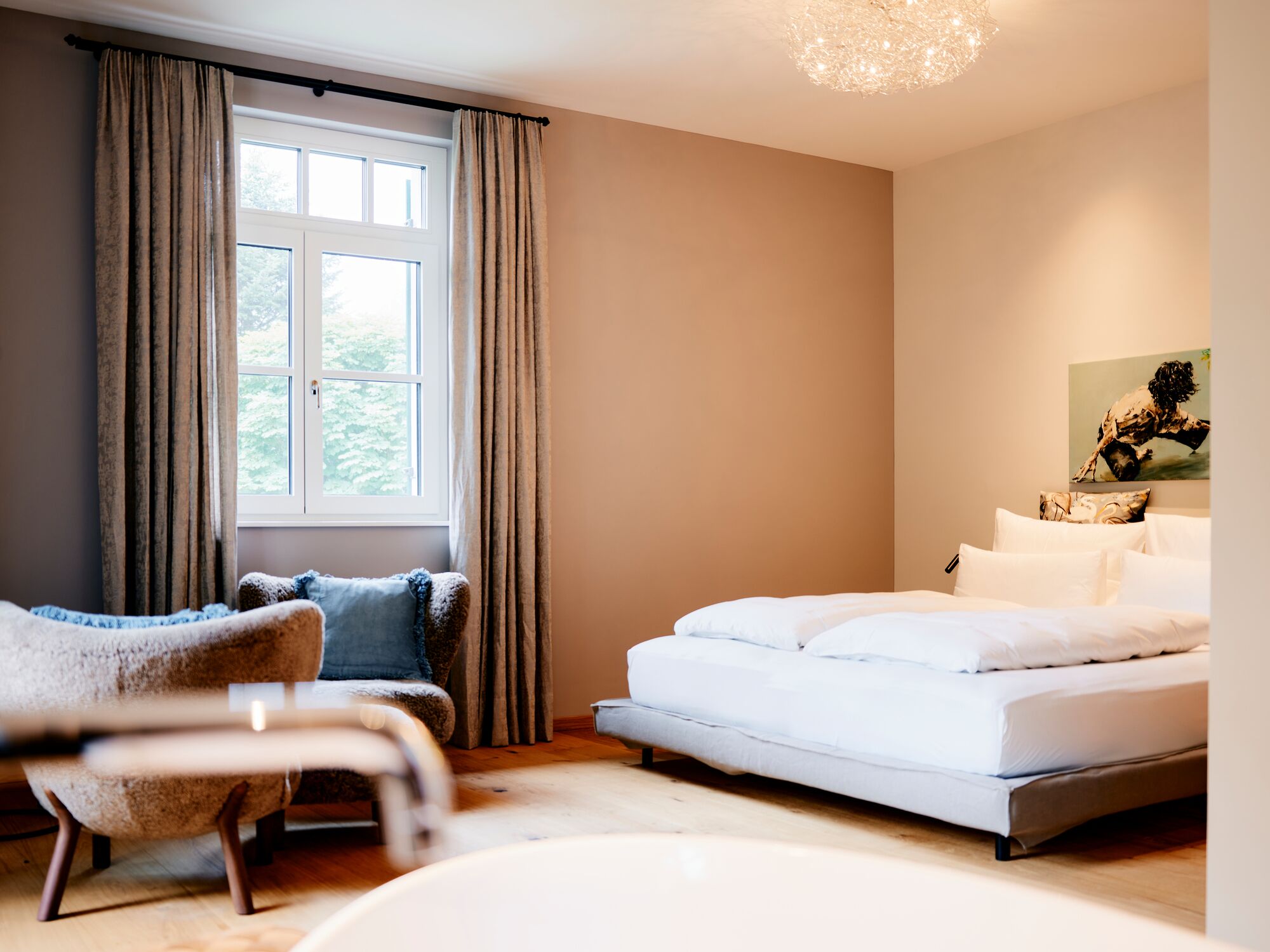 A bedroom with a bed, a chair, a window with curtains, and a painting on the wall.