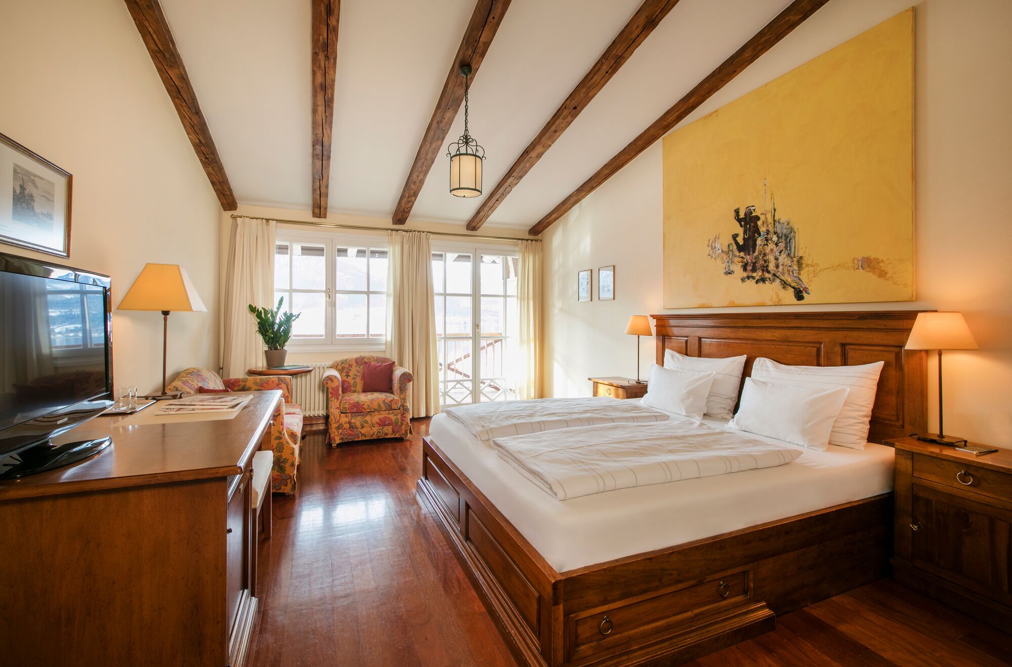 A hotel room with a wooden bed, a desk with a lamp, a chair, and a television.