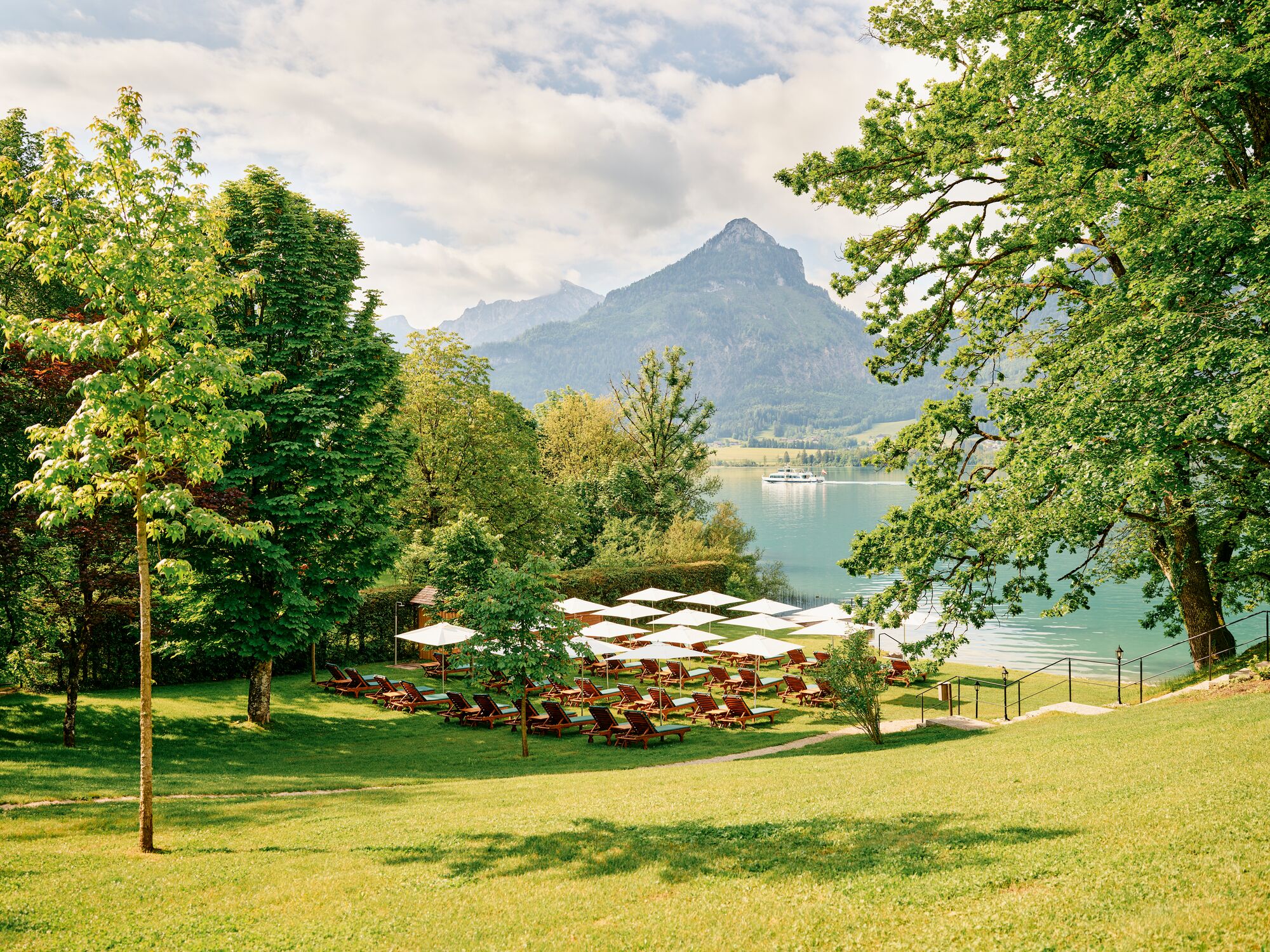 Un resort junto al lago con tumbonas y sombrillas, una montaña al fondo y cielo despejado.