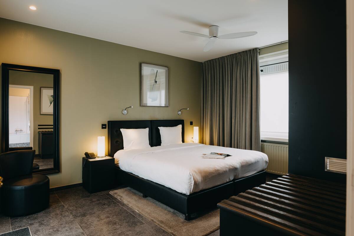 A hotel room with a large bed, bedside lamps, a ceiling fan, a large mirror, and a window with curtains.