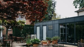 An outdoor patio area with tables and chairs, potted plants, and a modern building with large windows.