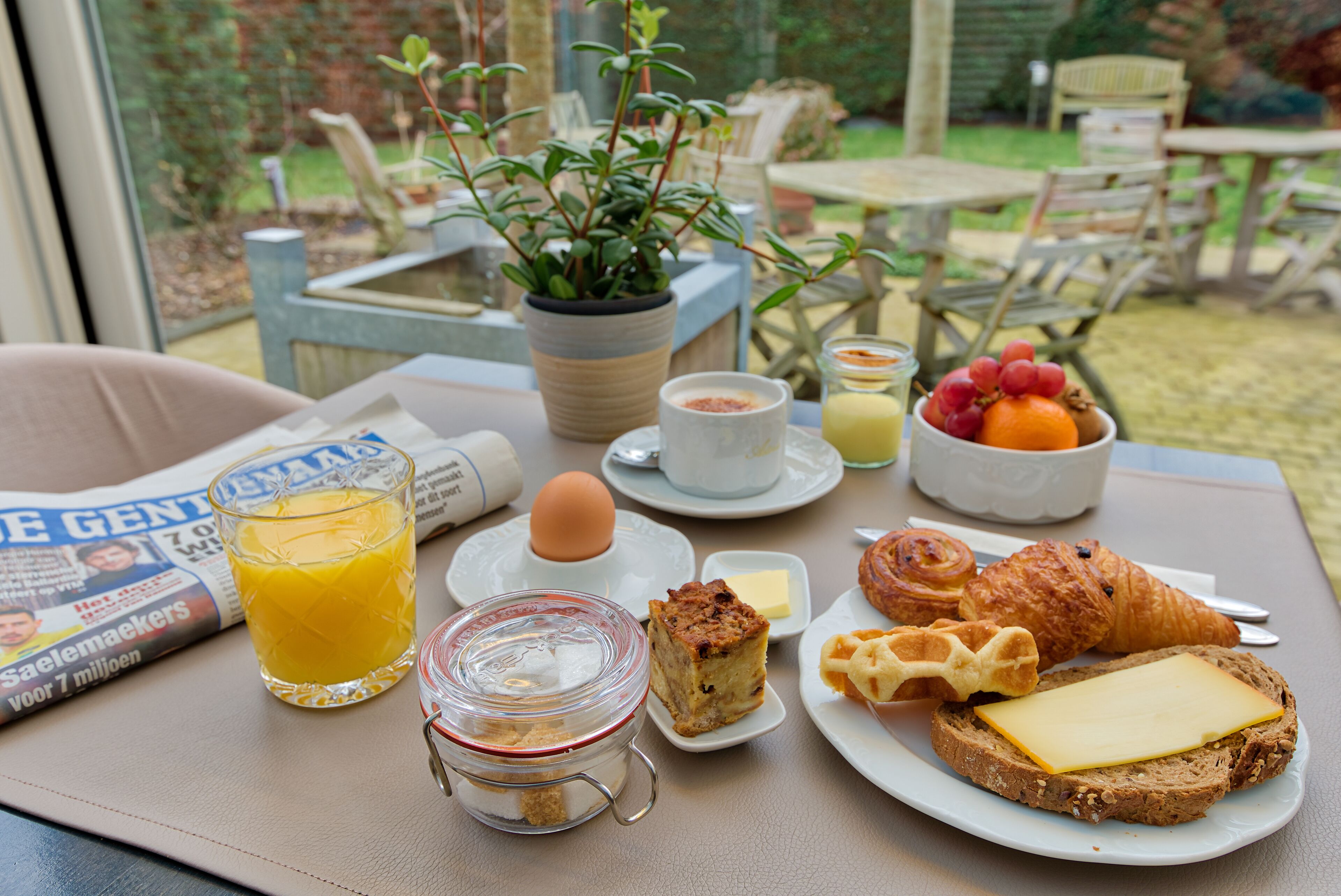 A breakfast table with croissants, cheese, fruit, a glass of orange juice, a newspaper, and a potted plant.