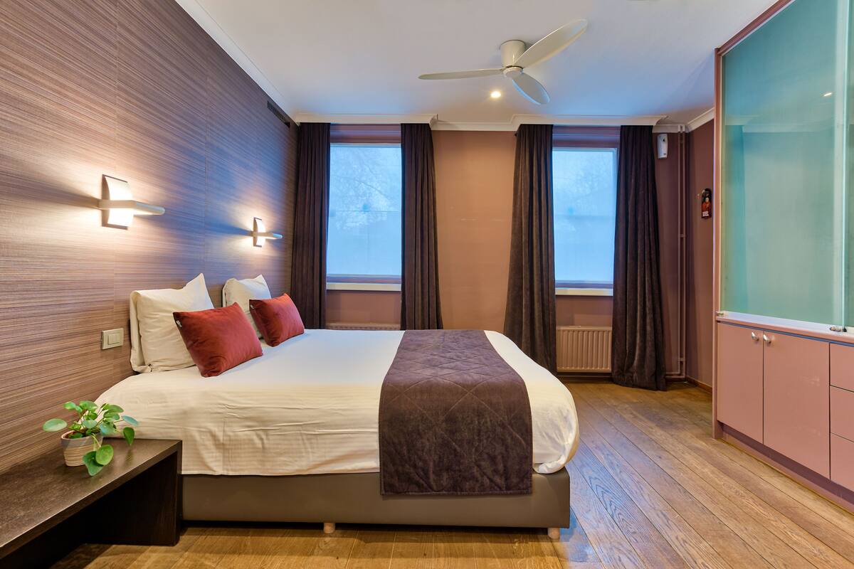 A modern bedroom with a large bed, a ceiling fan, and a view of the outdoors through two windows.
