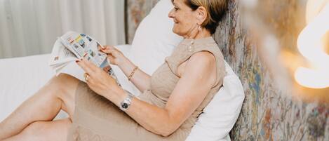 A person sitting on a bed reading a magazine in a room with a floral wallpaper and a white pillow.