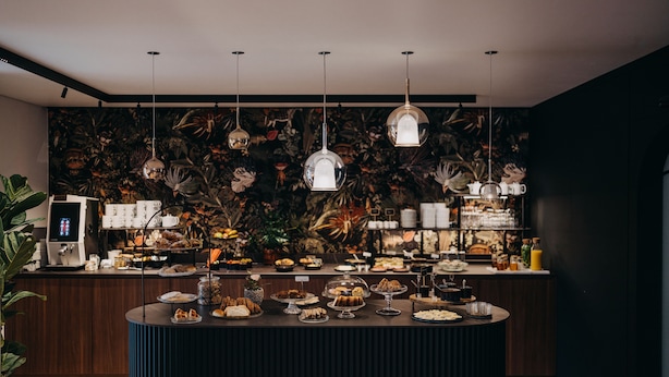 A buffet with various food items and a coffee station, set against a dark wall with a tropical mural.