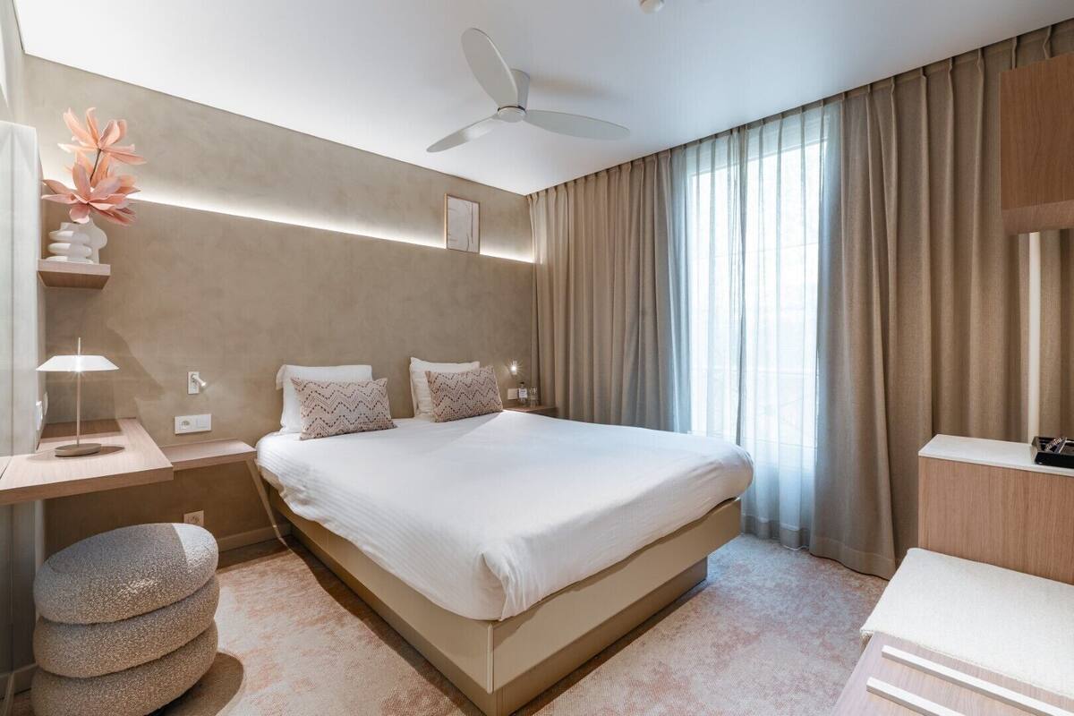 A modern hotel room with a large bed, a desk with a lamp, a small seating area, and a ceiling fan.