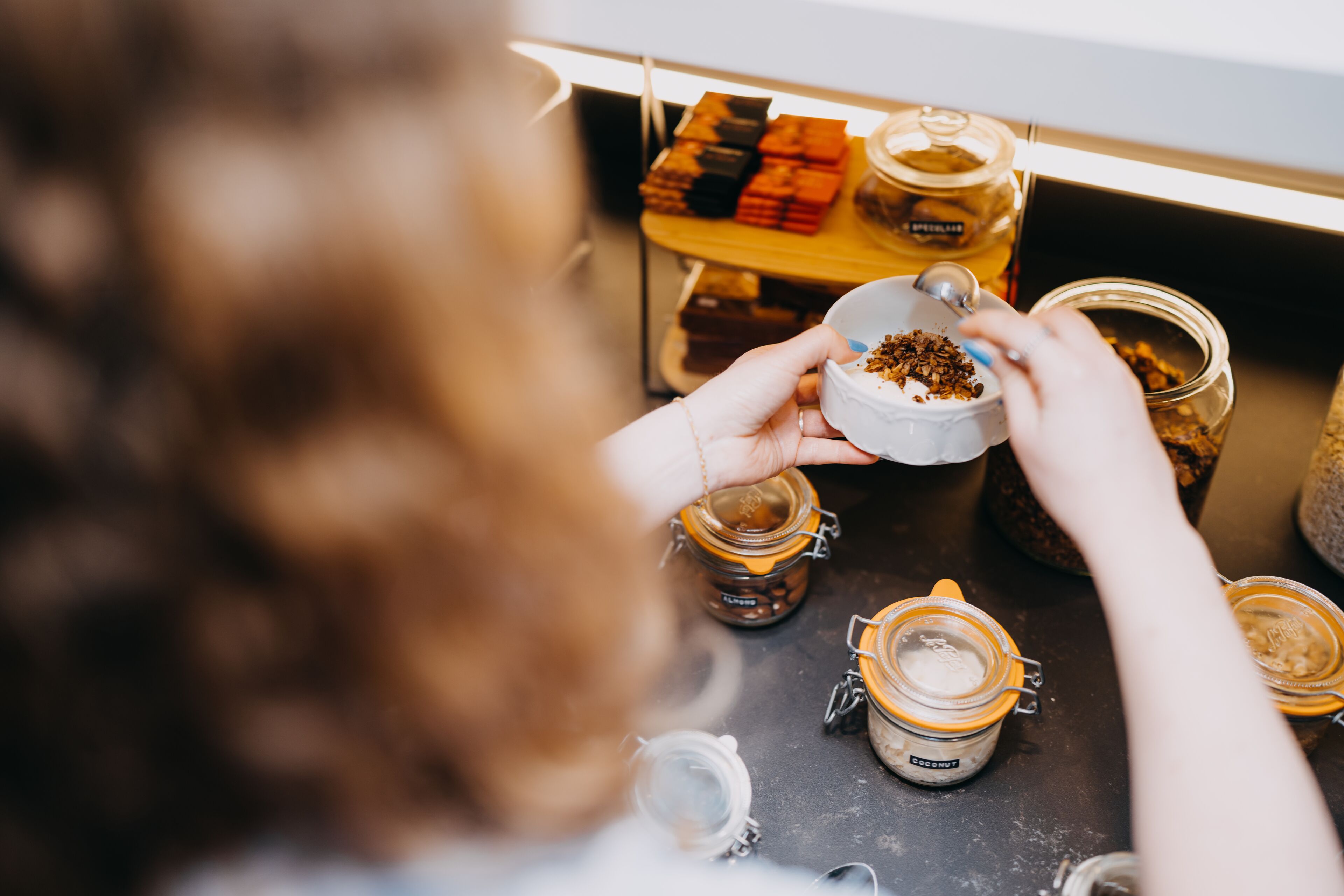 A person holding a small bowl with a spoon, surrounded by jars and containers on a table.