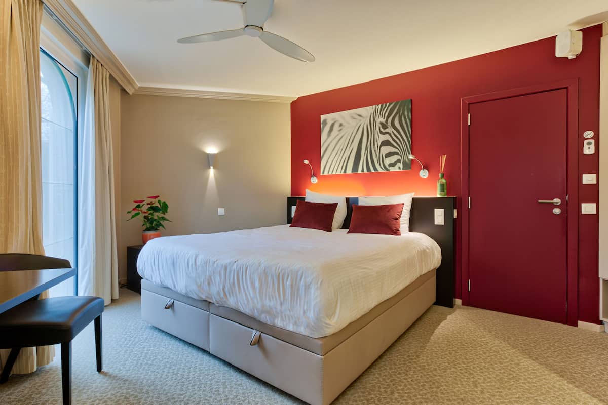 A hotel room with a bed, a desk, a chair, a ceiling fan, a red door, and a wall with a zebra print artwork.