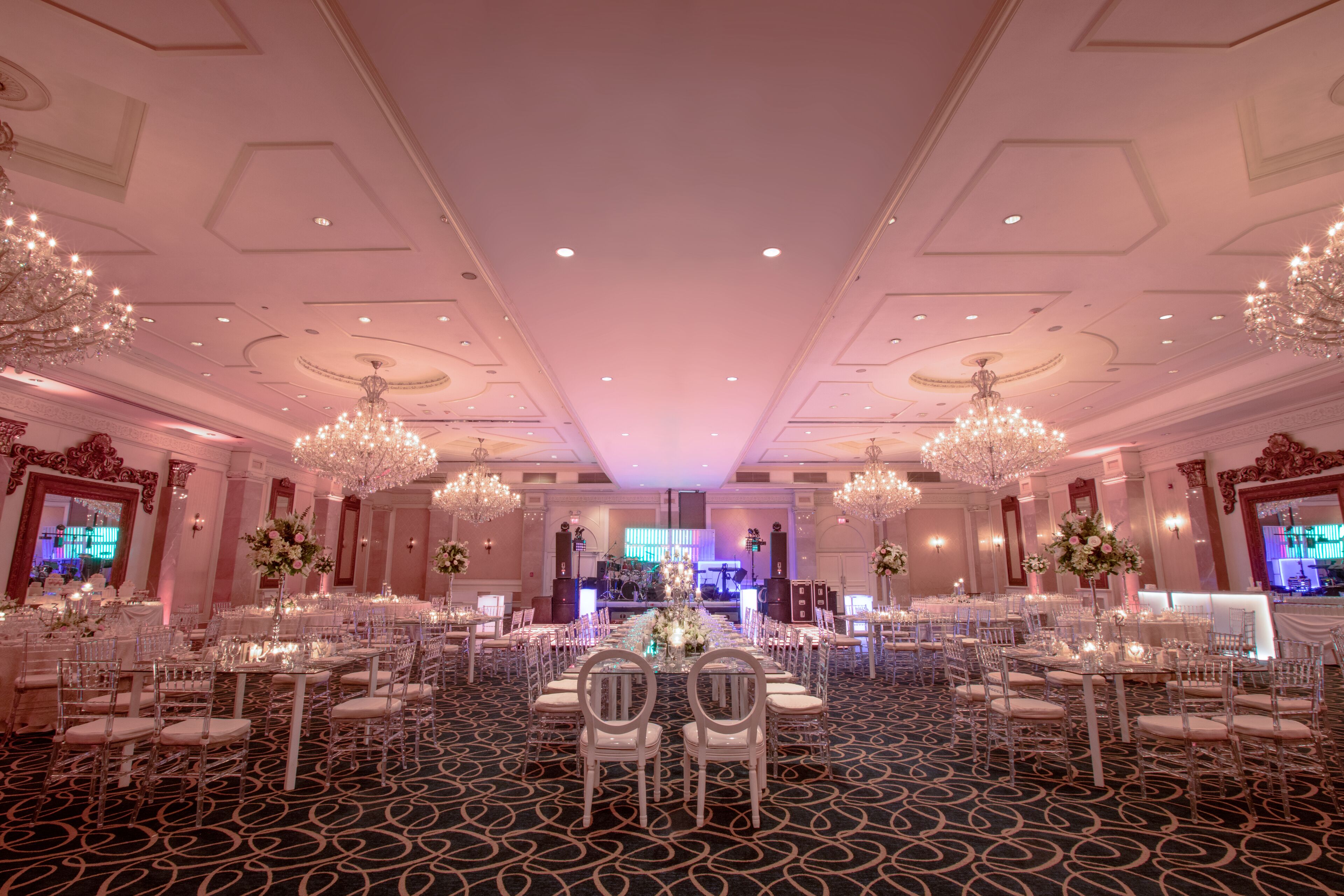 A banquet hall with round tables, chairs, chandeliers, and floral centerpieces.
