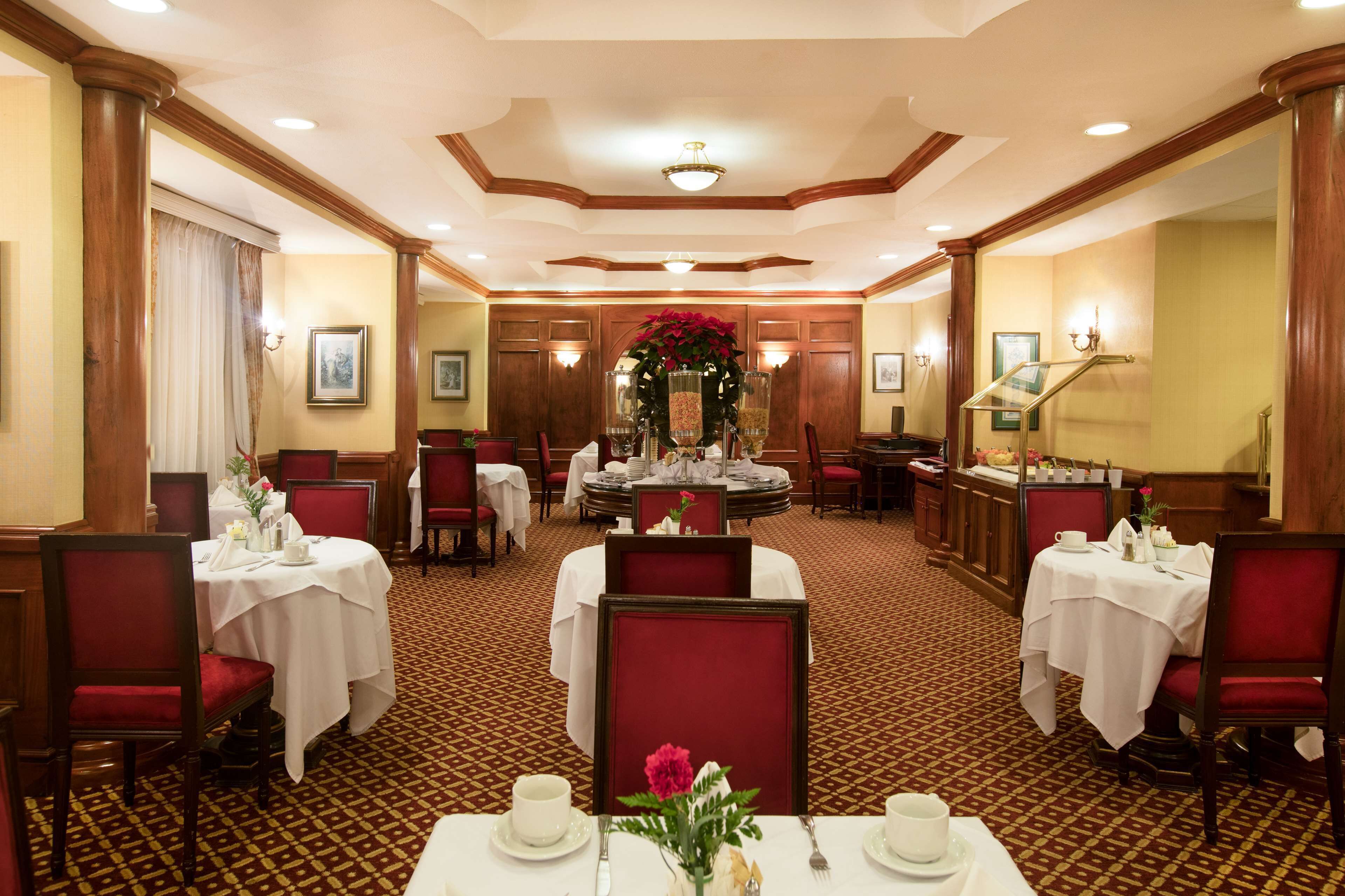 A hotel dining area with tables set for service, red upholstered chairs, and a central decorative piece.