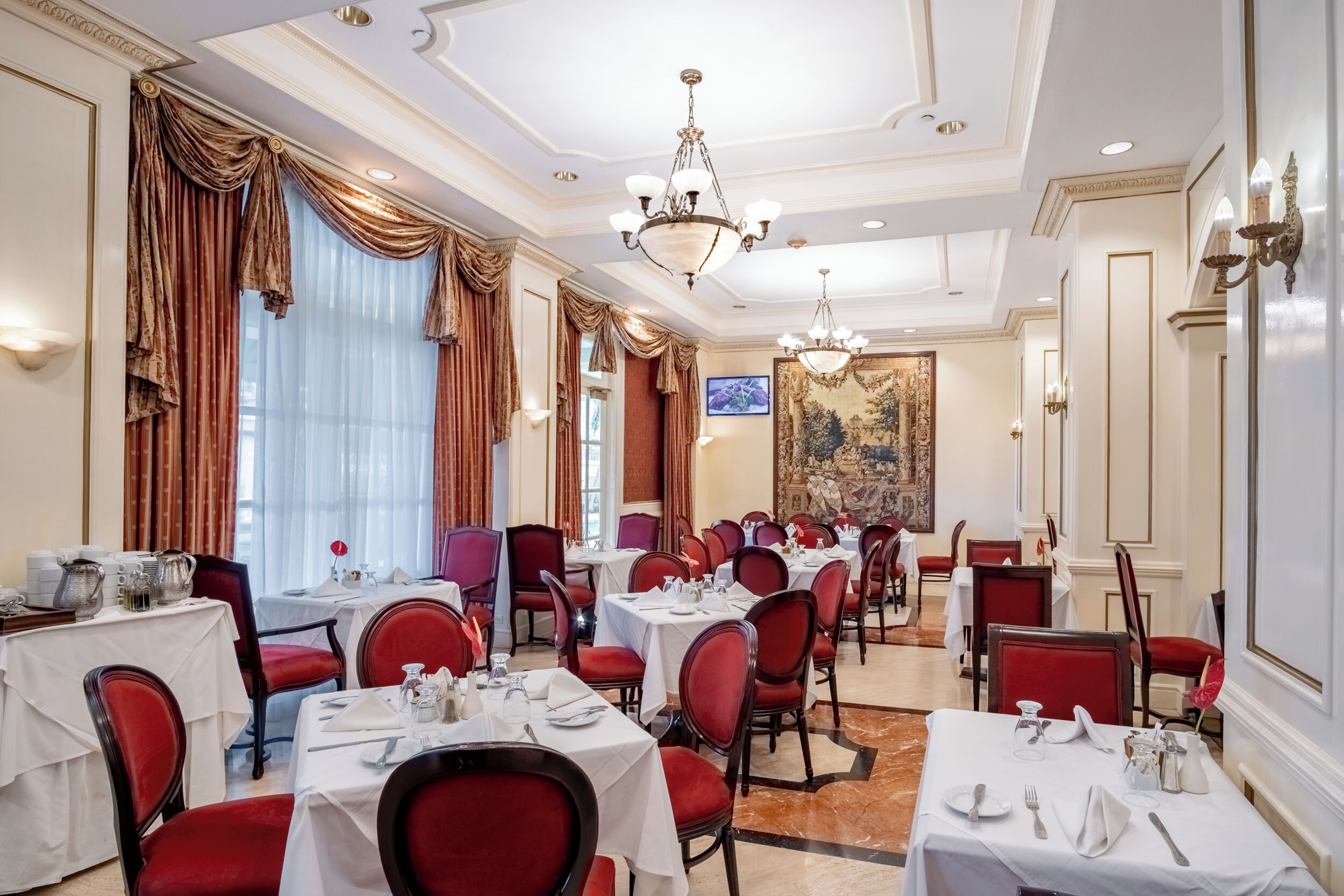 A formal dining room with tables set for service, red upholstered chairs, chandeliers, and a large painting on the wall.