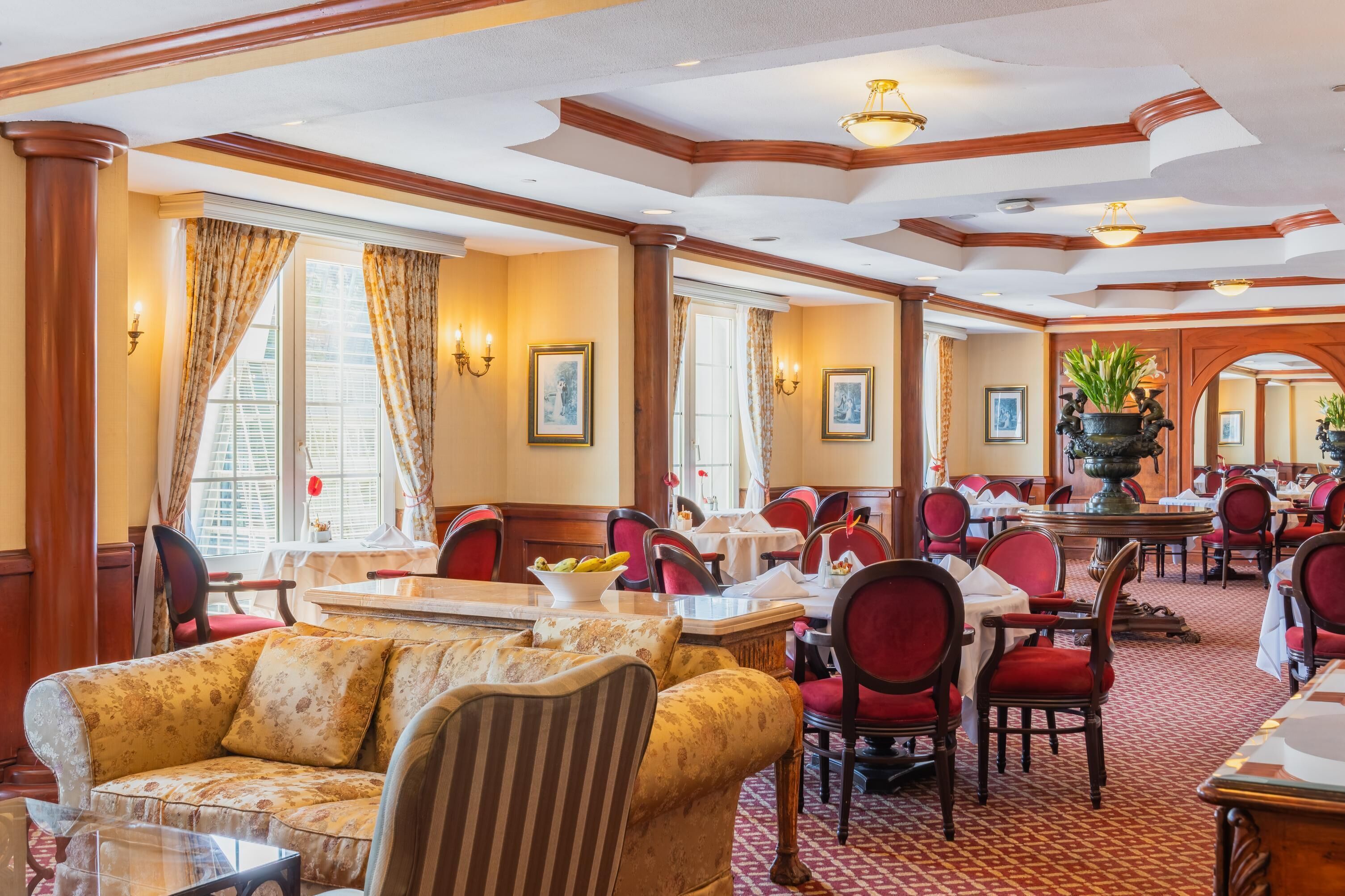 A hotel dining area with multiple tables and chairs, a sofa, and a large vase with plants.