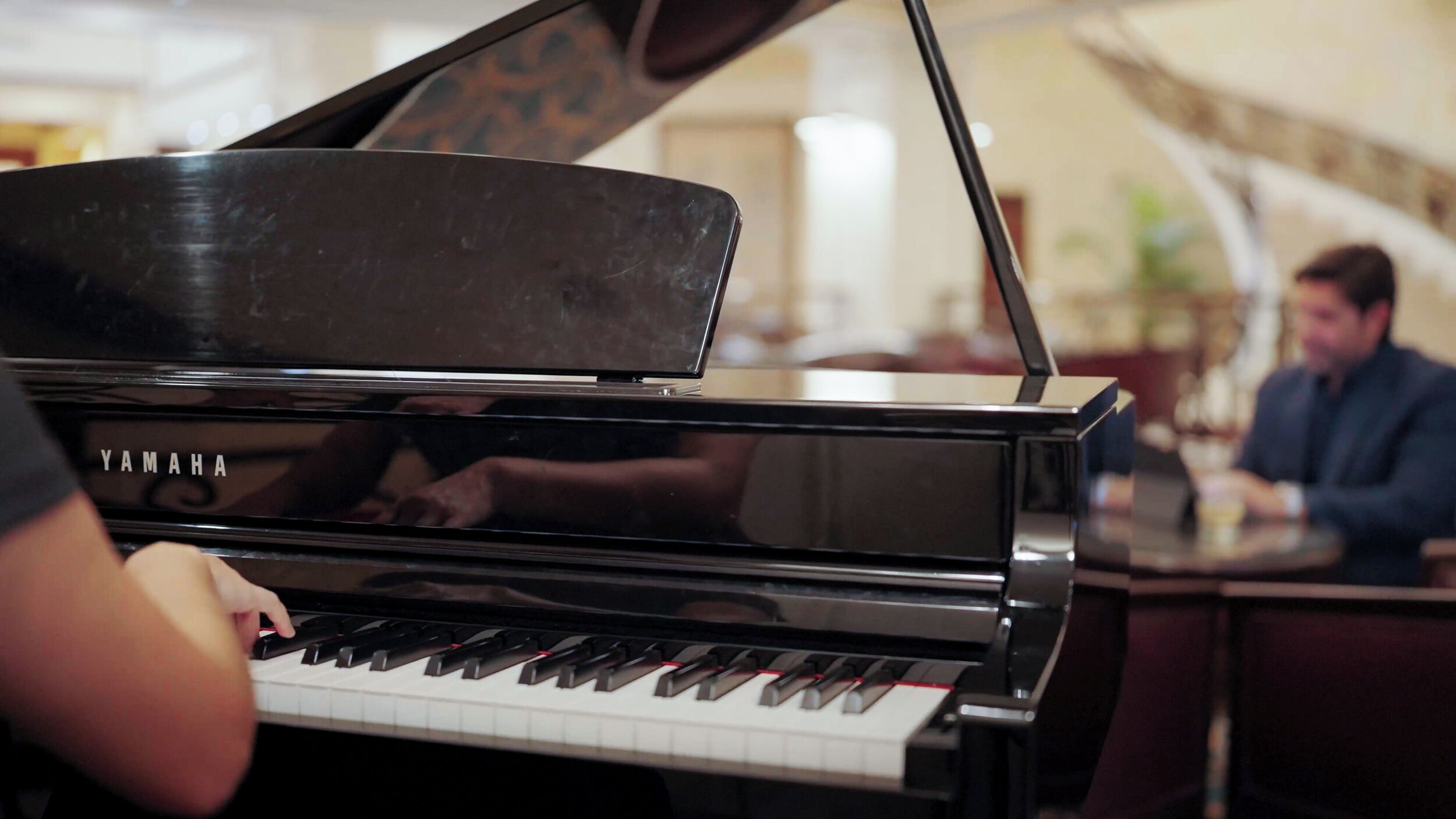 Una persona tocando un piano de cola Yamaha en una habitación con zona de comedor y otra persona sentada a una mesa al fondo.