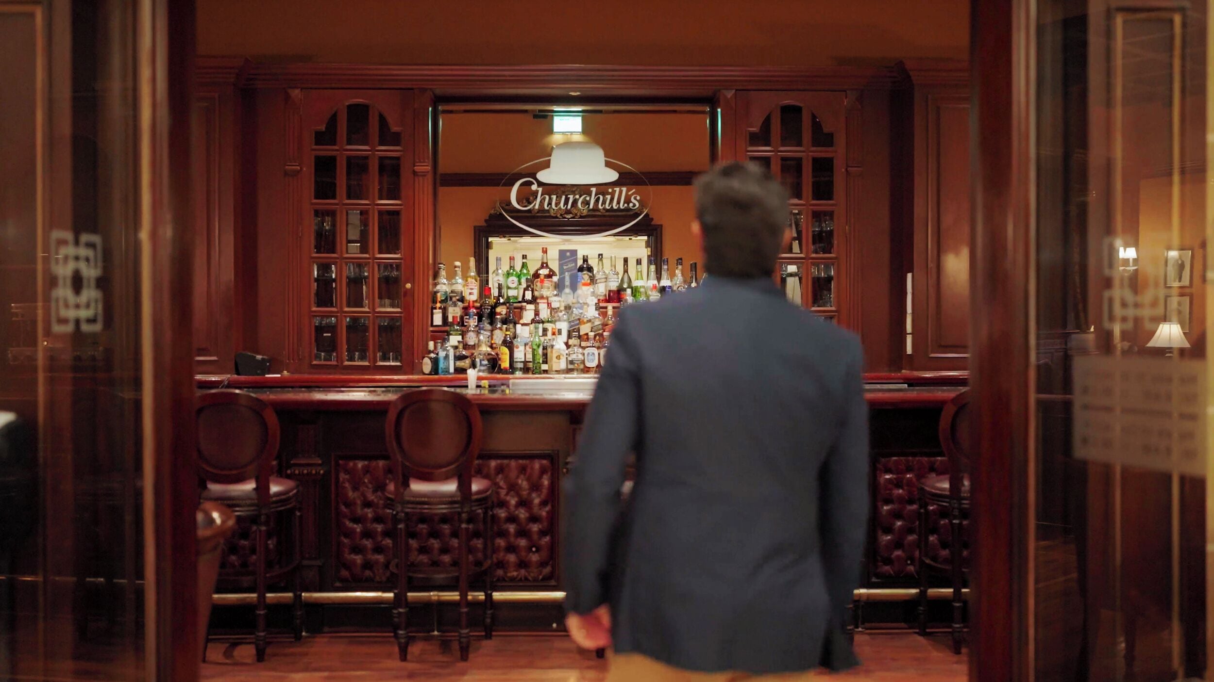 A man walking towards a bar with a sign reading 'Churchill's' and a well-stocked bar area.