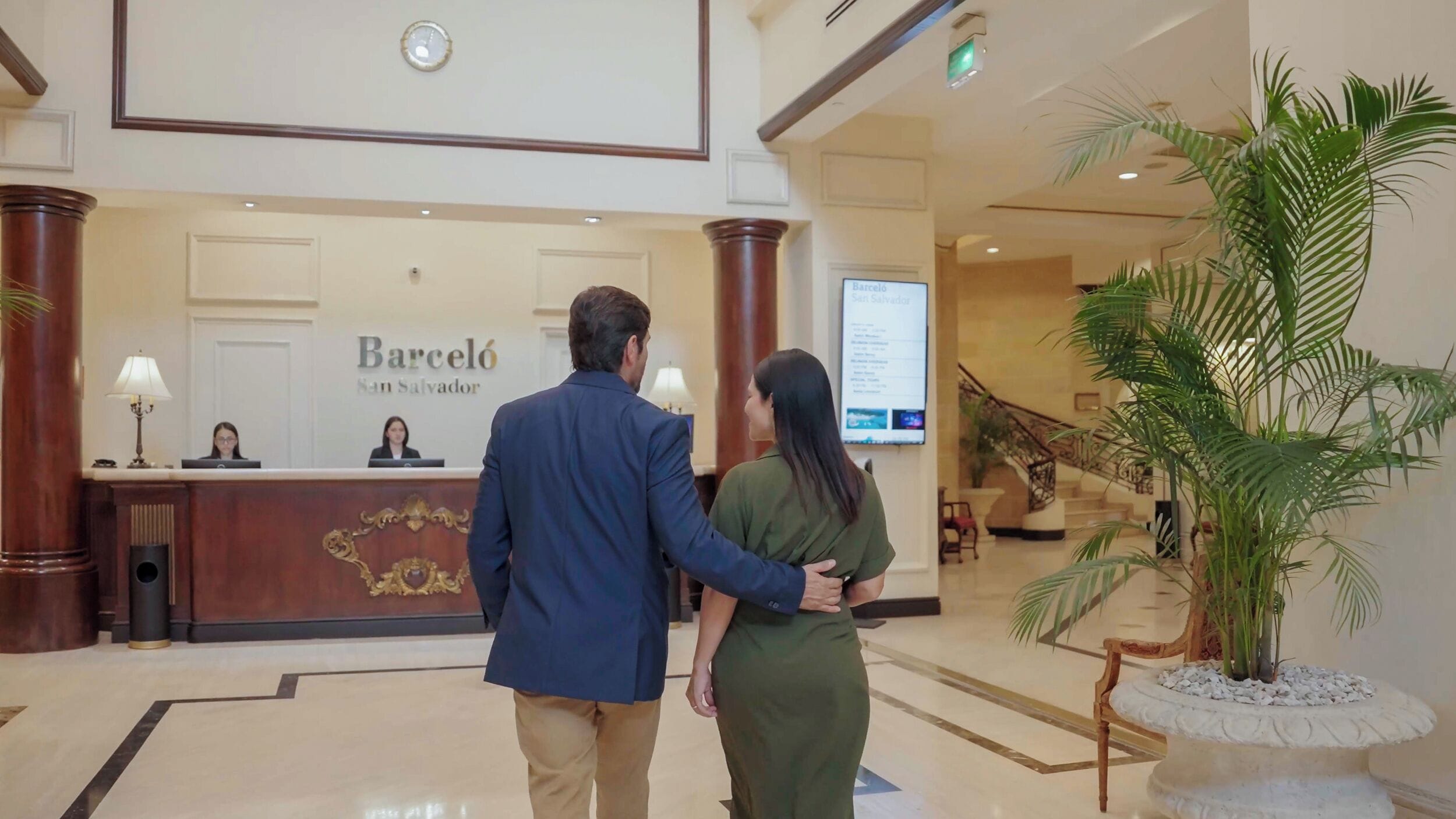 A man and woman walking towards a reception desk in a hotel lobby with a staircase and a large potted plant.
