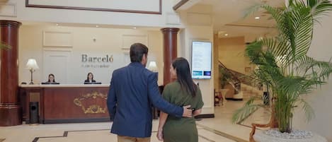Un hombre y una mujer caminando hacia la recepción en el vestíbulo de un hotel, con una escalera y una planta grande en maceta.