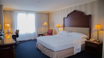 Een hotelkamer met een groot bed, twee nachtkastjes met lampen, een bureau met stoel, en een raam met gordijnen.