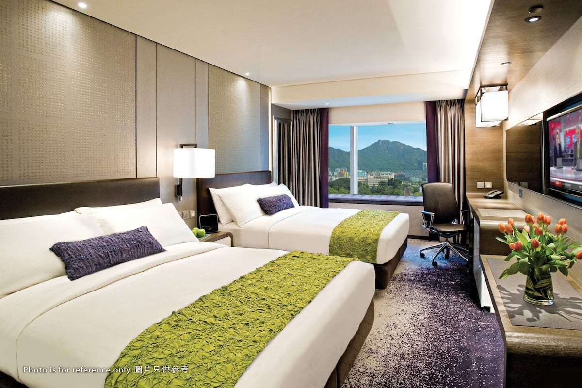 A hotel room with two beds, a desk, a TV, and a view of mountains.
