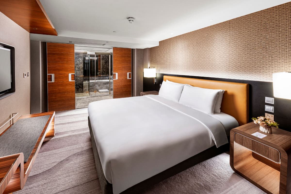 A modern hotel room with a large bed, a desk, and a glass door leading to a balcony.