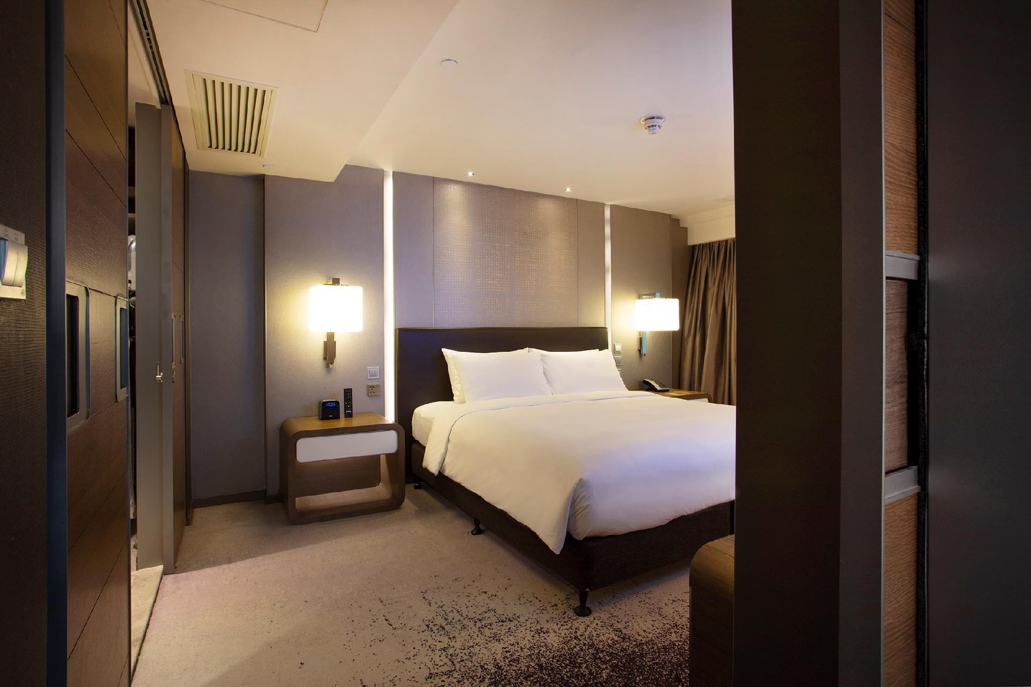 A hotel room with a large bed, bedside lamps, a nightstand, and a wall-mounted heater.